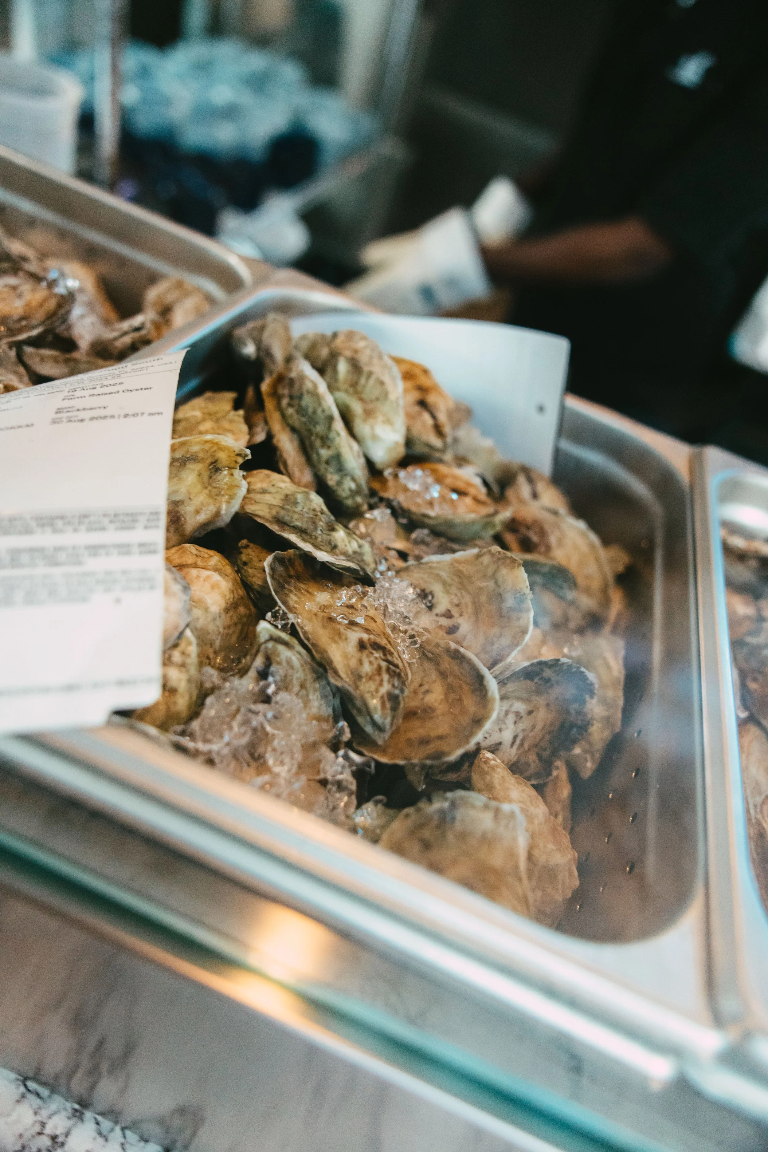 Fresh oysters on ice in a seafood display tray, with a customer in the background.