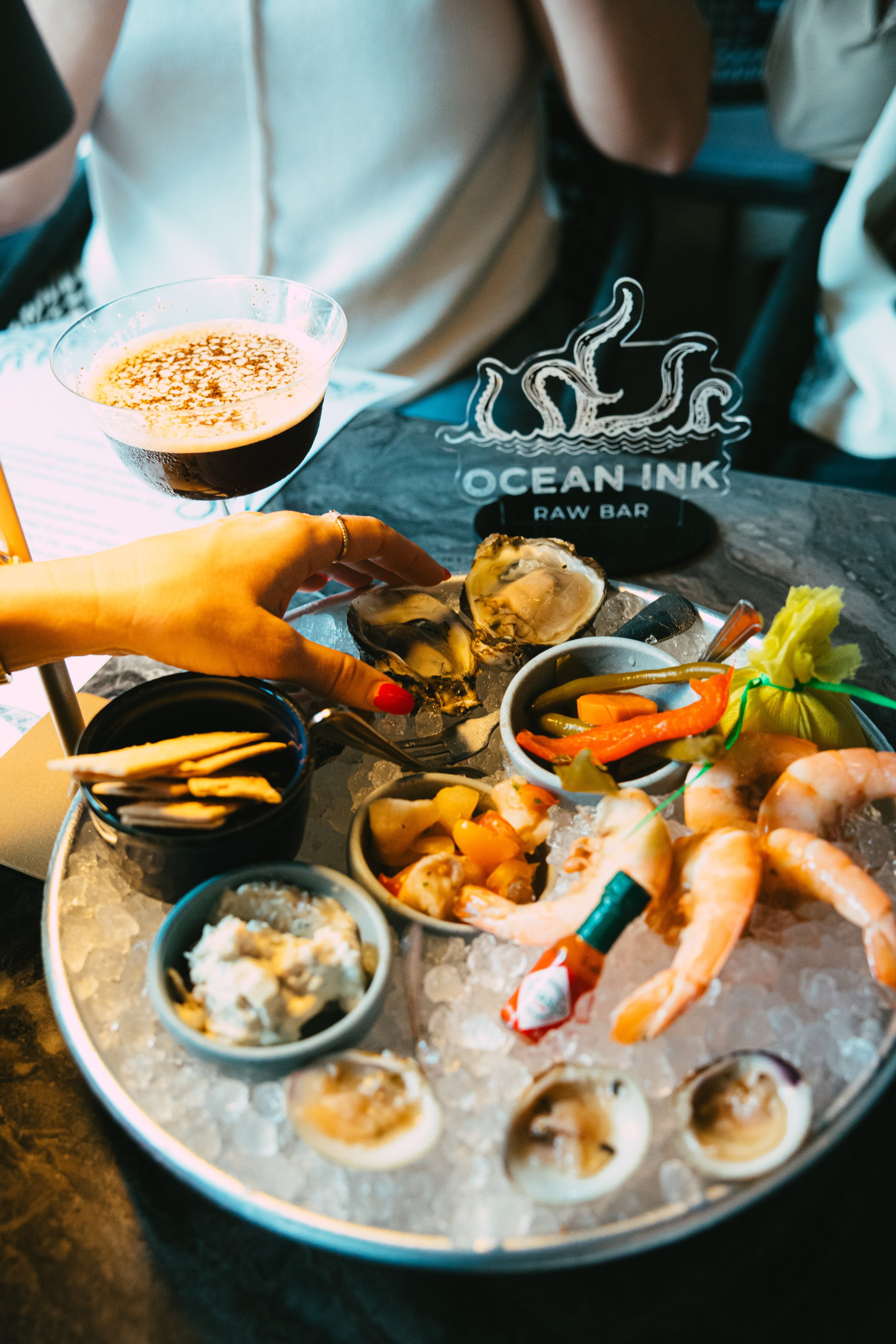 Seafood platter with oysters, shrimp, and vegetables on ice, alongside a glass of dark beer at Ocean Ink Raw Bar.