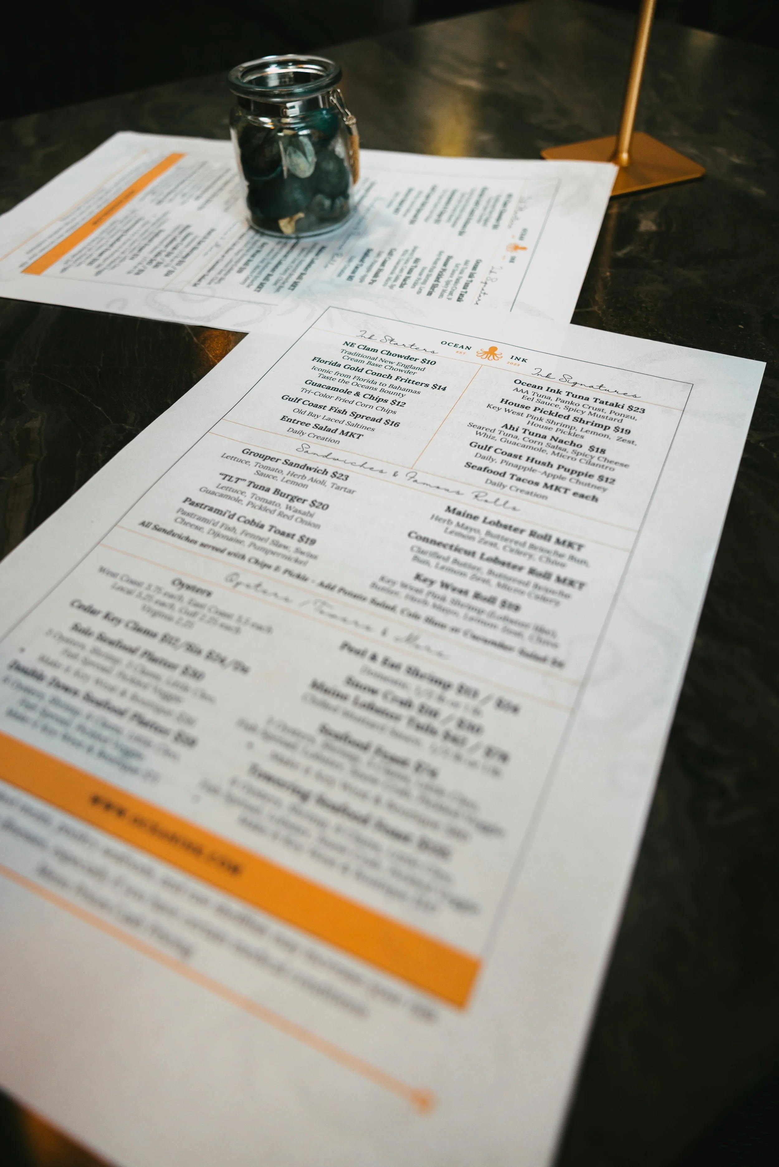 Restaurant menu lying on a dark table with a small jar filled with coins and a gold-colored pen holder in the background.