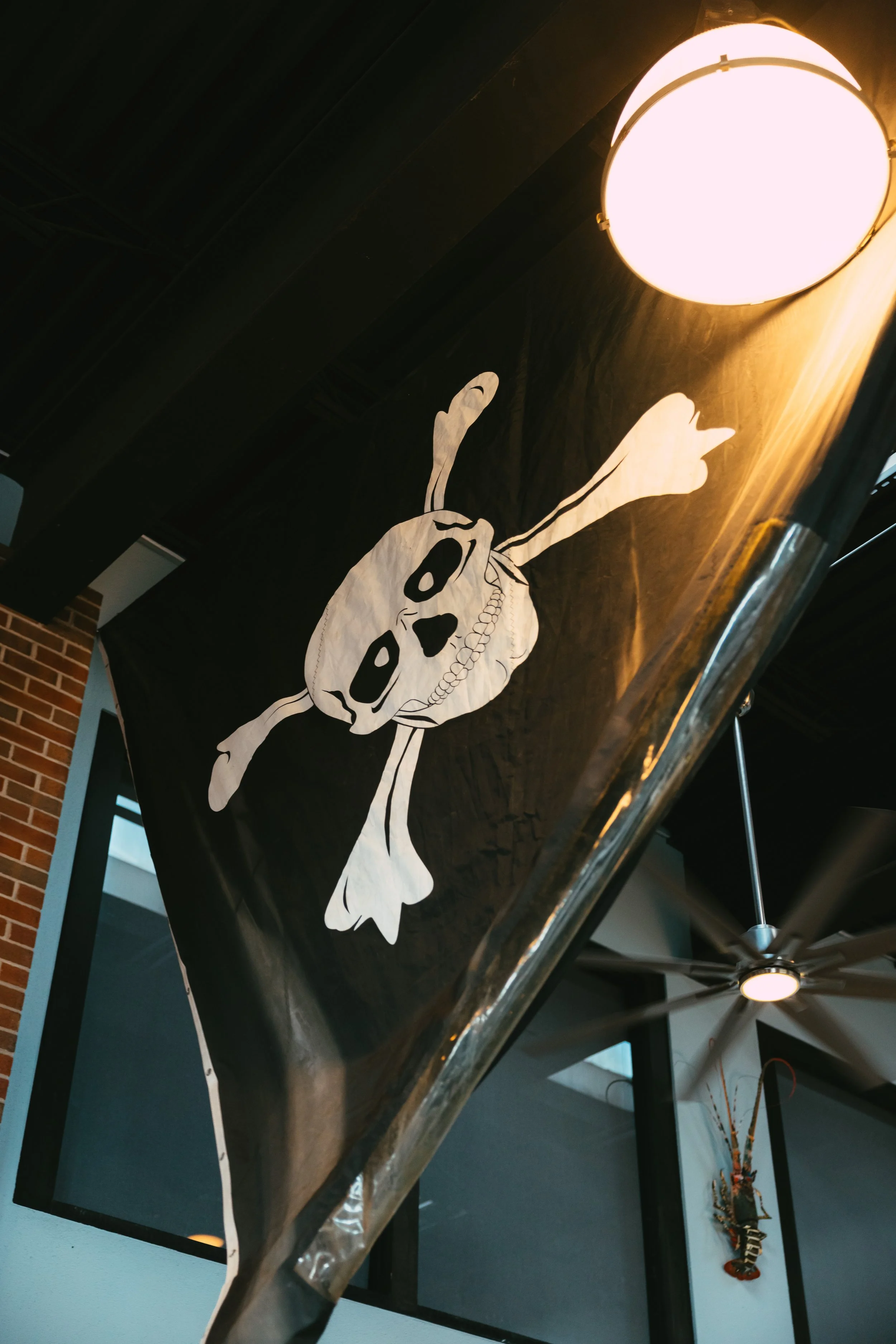 Black flag with a white skeleton skull and crossbones design hanging from ceiling near a bright round light fixture in a restaurant or bar interior.