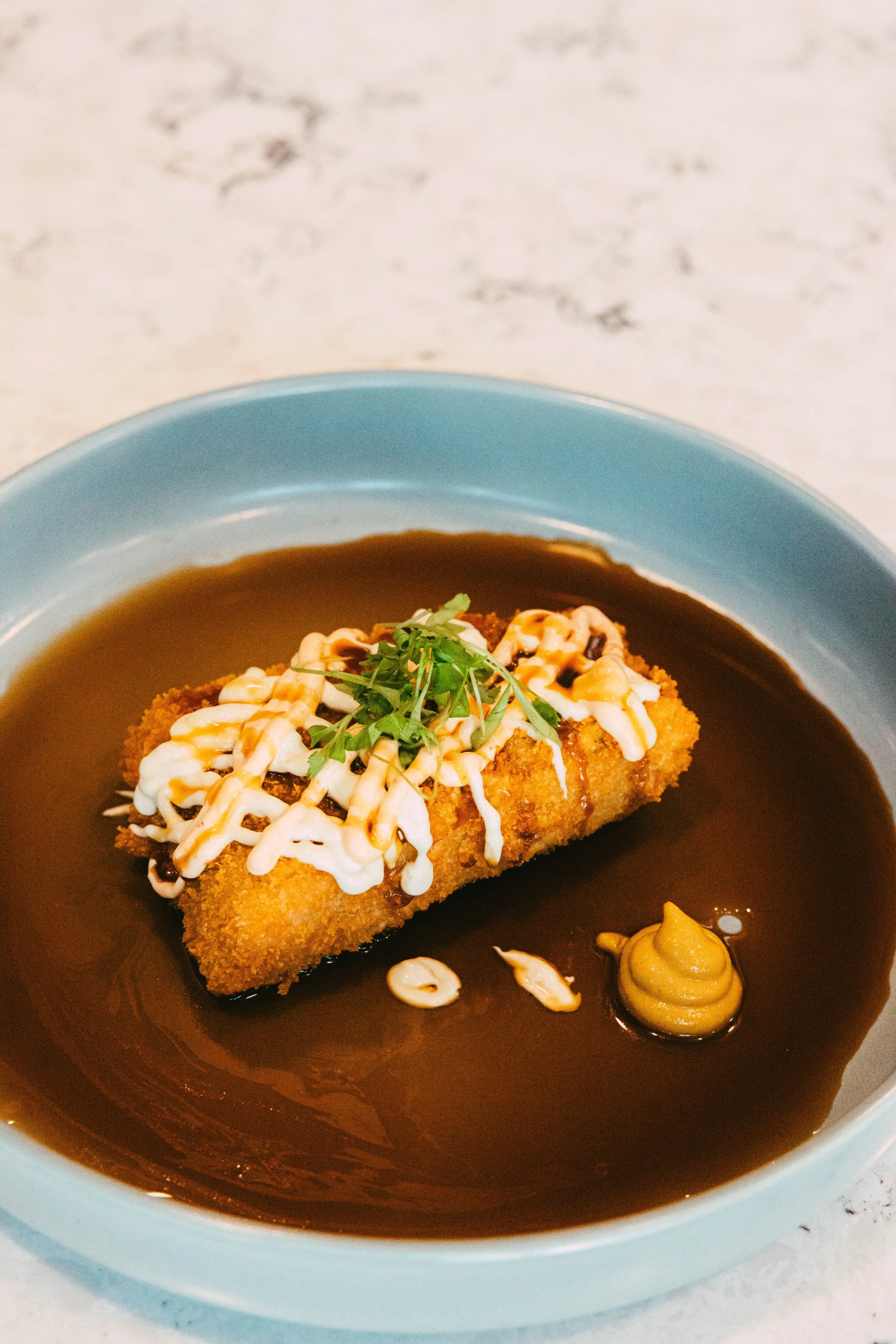 Fried fish fillet topped with mayonnaise, garnished with herbs, served in dark gravy with mustard on a blue plate.
