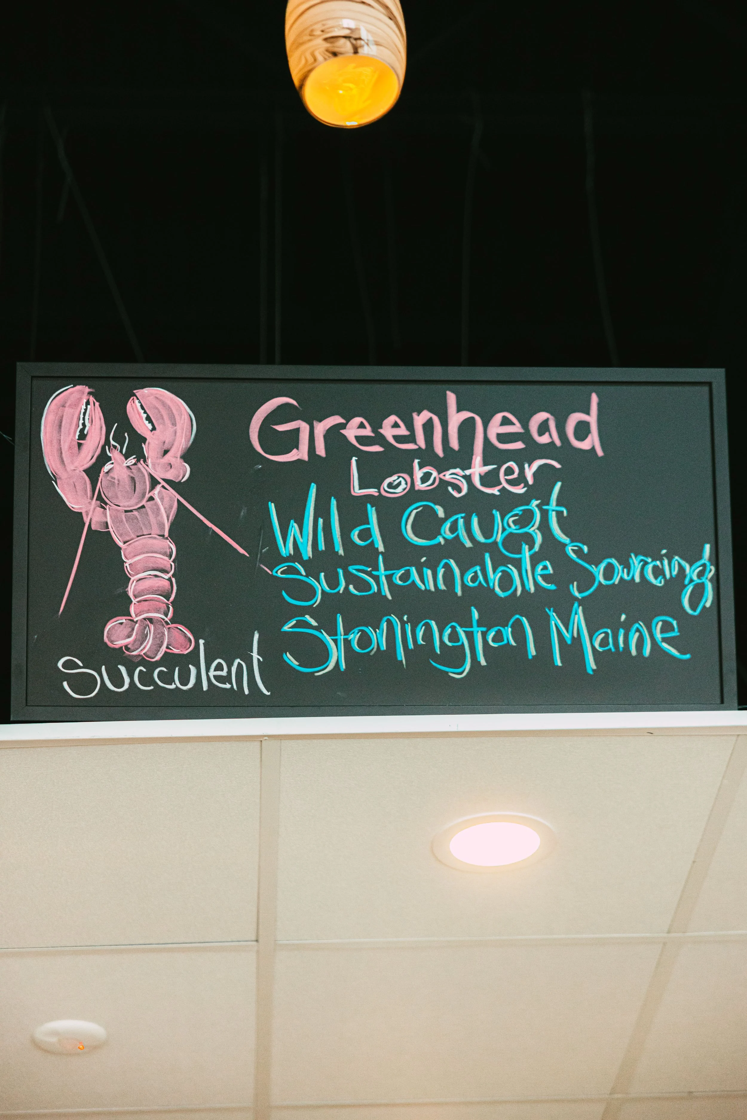 A blackboard sign displaying a pink lobster drawing and the words "Greenhead Lobster, Wild Caught, Sustainable Sourcing, Stonington Maine, Succulent" in pink, blue, and white chalk.