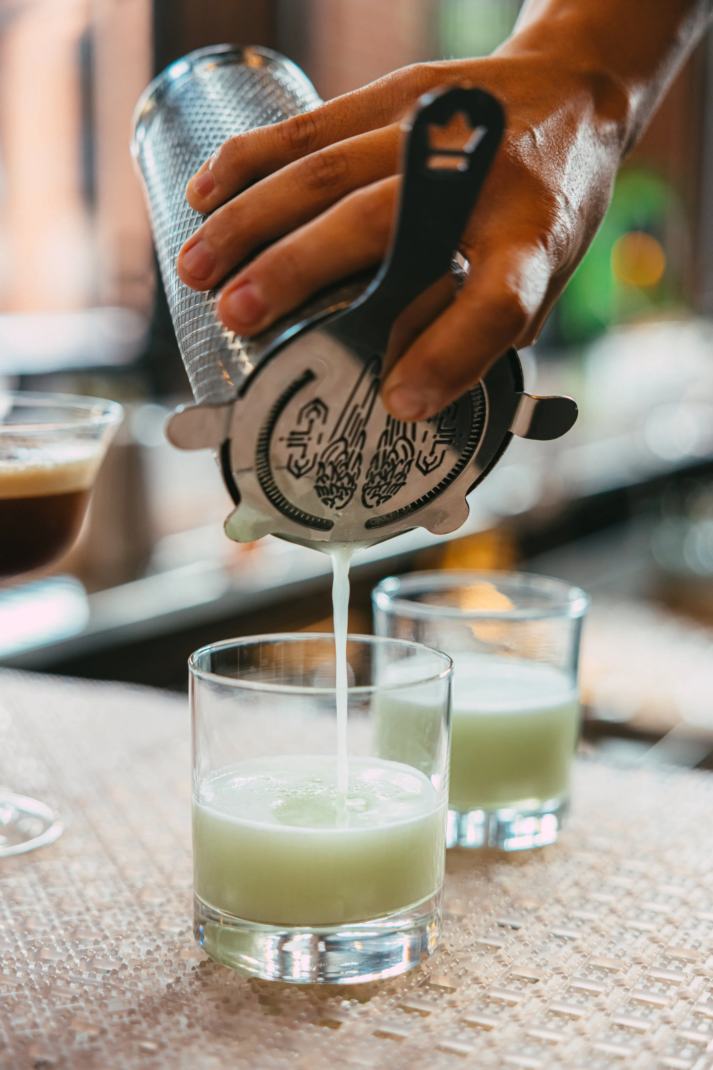 Person pouring a green cocktail from a cocktail shaker into a glass, with another partially filled glass of the same drink on a textured surface.