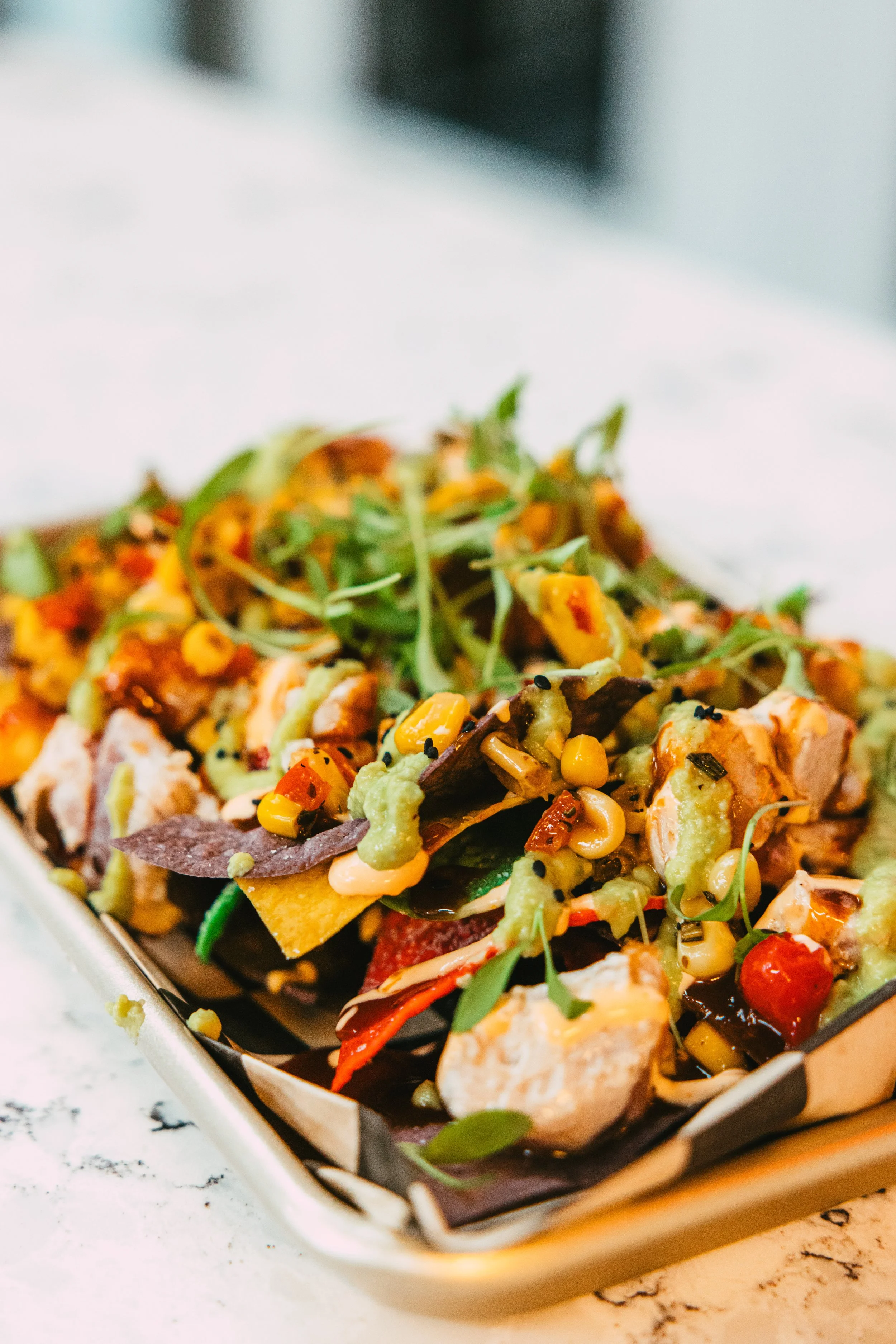 A close-up of a dish with mixed vegetables, crispy chips, greens, and toppings in a rectangular tray