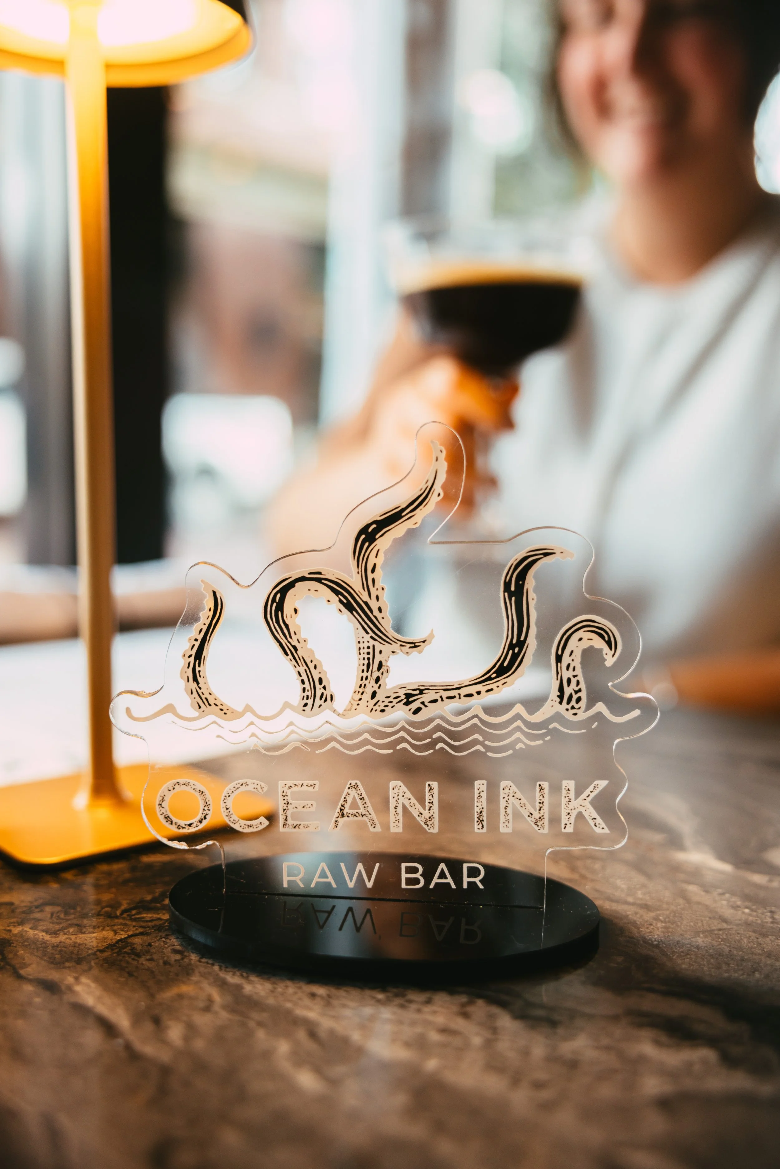 A decorative sign for Ocean Ink Raw Bar with an illustration of an octopus and the text "OCEAN INK RAW BAR" on a table. In the background, a woman smiling, holding a glass of dark drink.