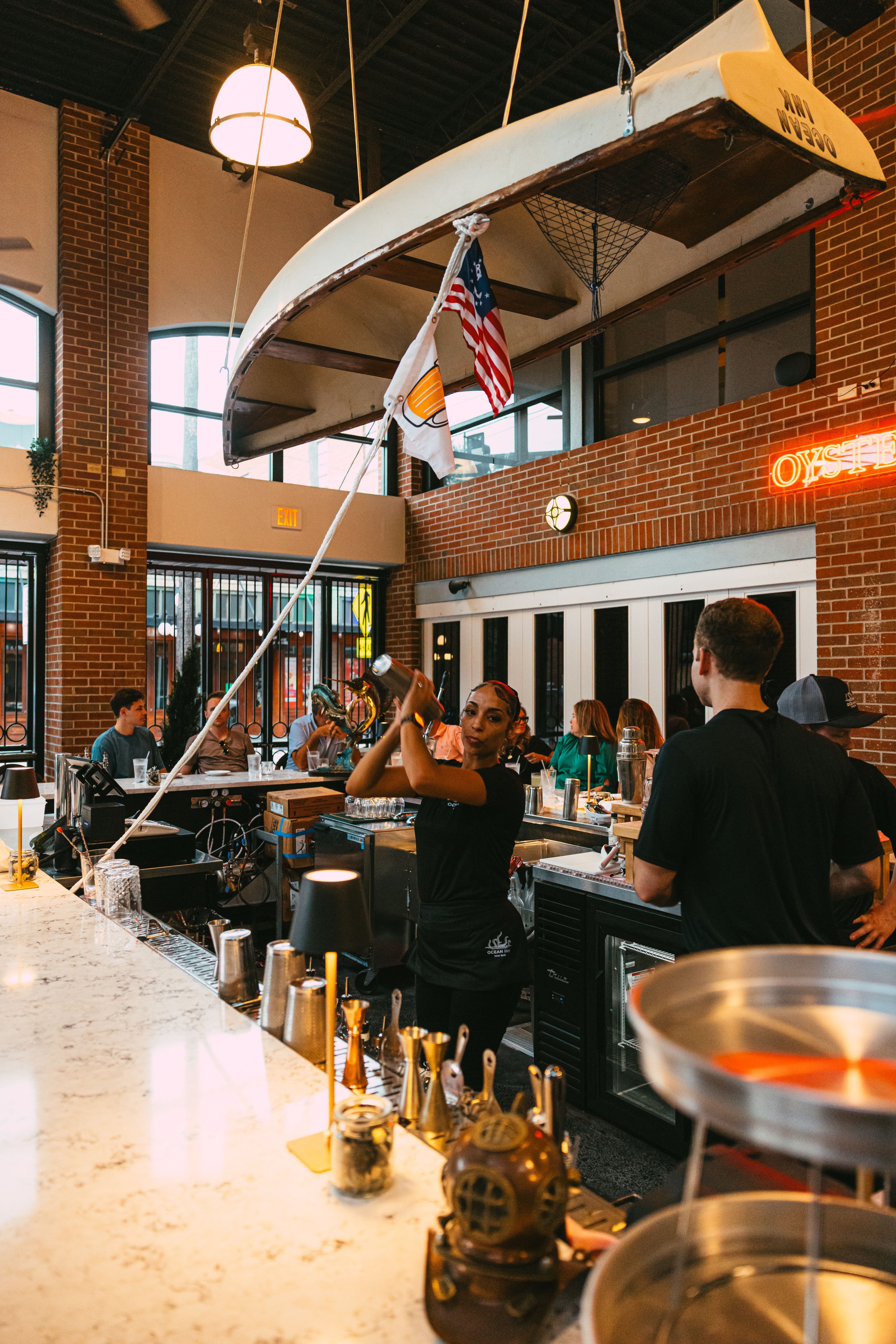 A bartender shaking a cocktail in a bar with a large paddleboard hanging from the ceiling and American flags.