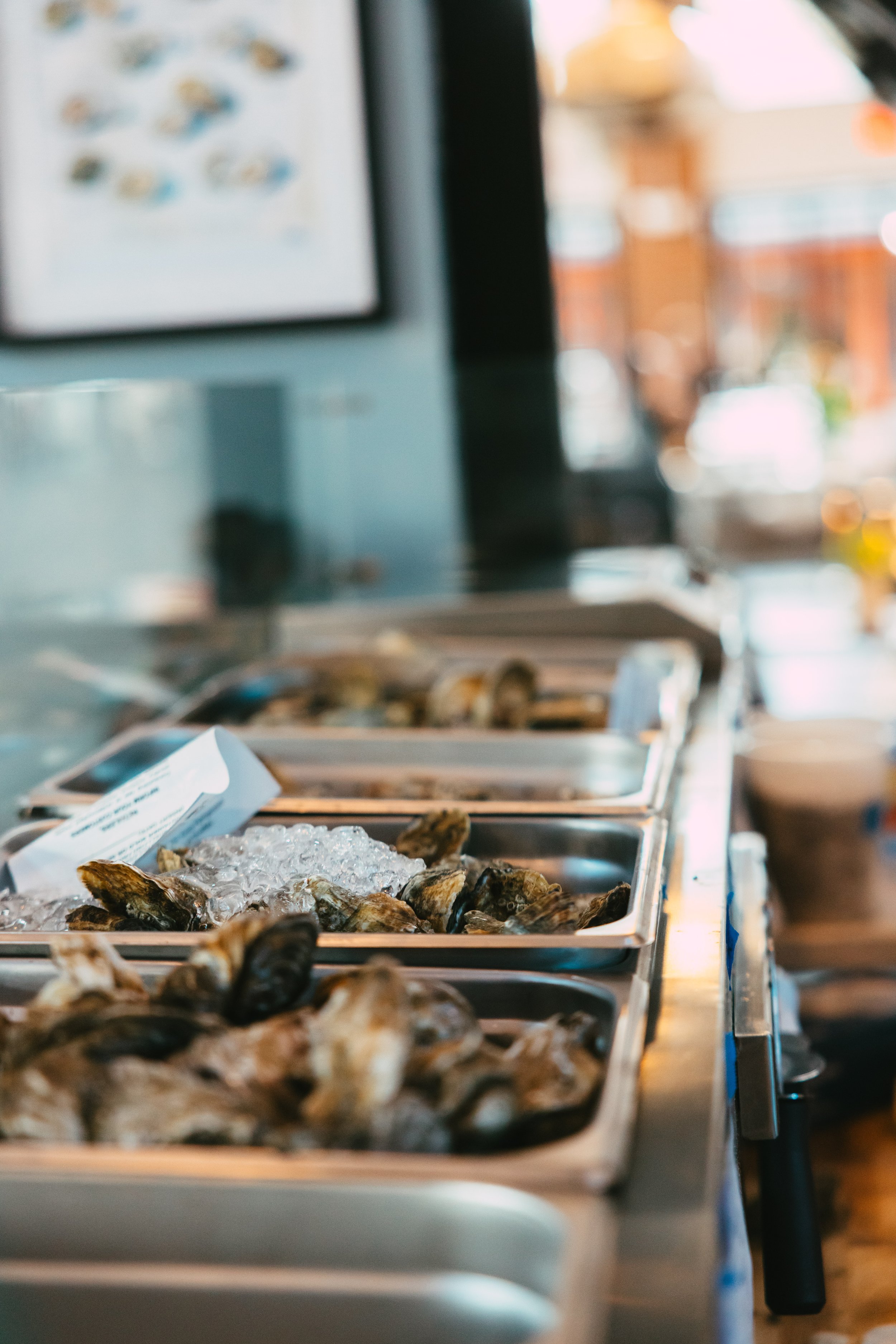 Display of fresh oysters on ice at a seafood market or restaurant, with seafood trays and a blurred background.