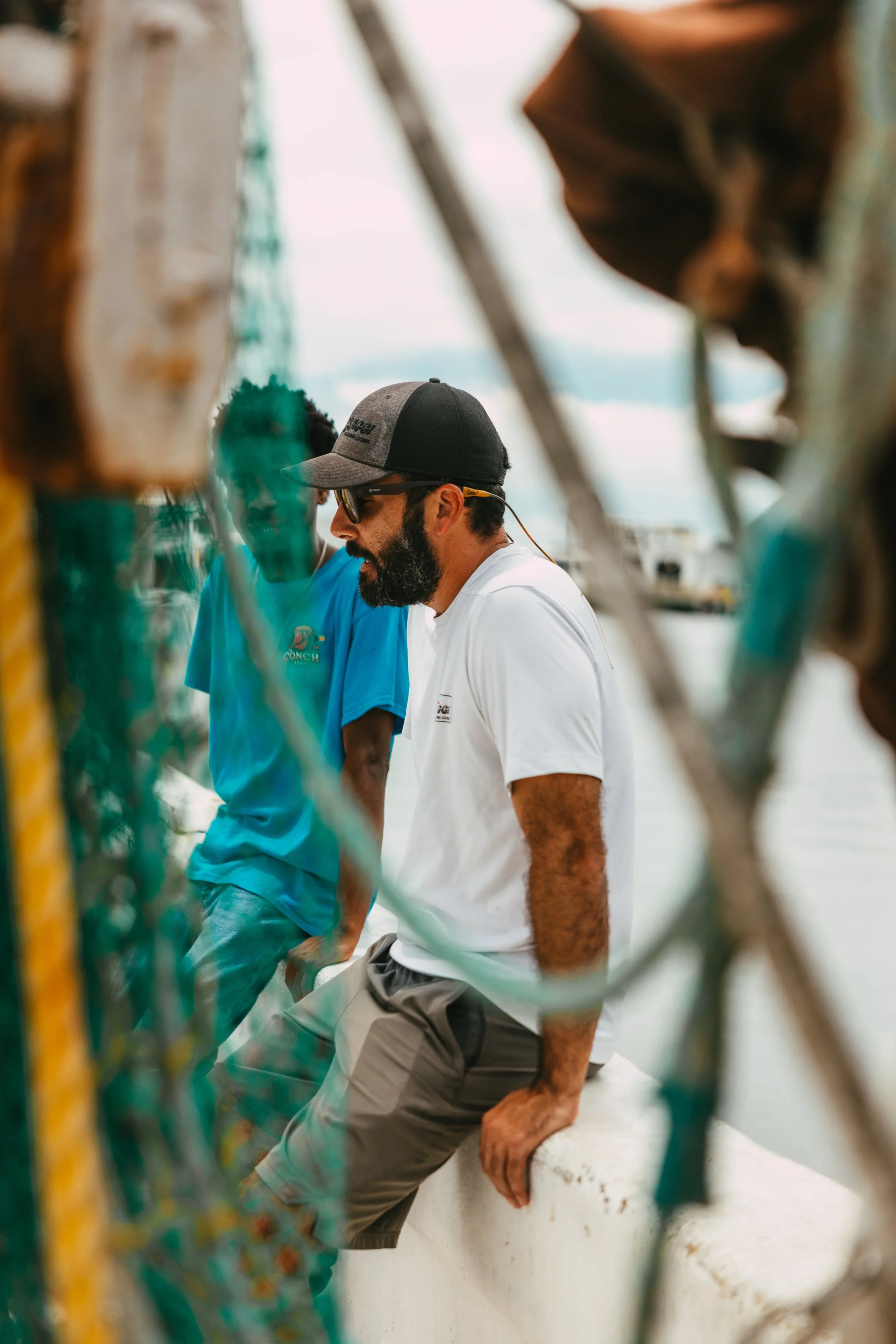 Two men sitting on a dock by the water, framed by fishing nets and ropes, engaged in conversation.
