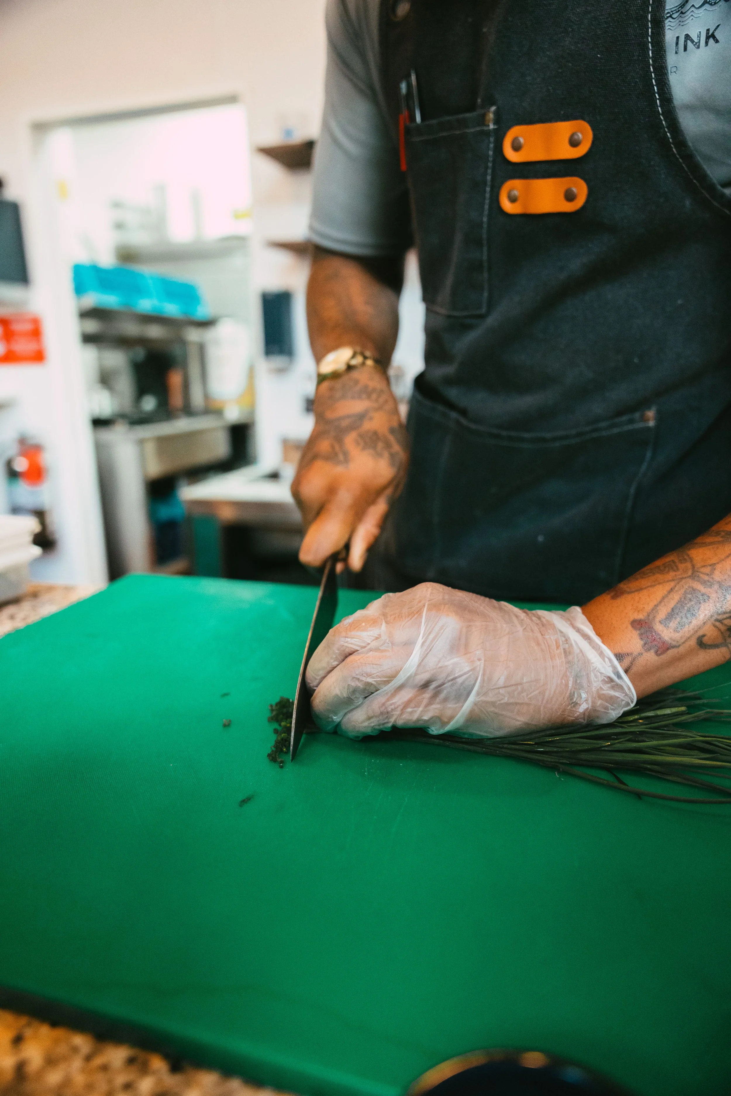 A person wearing gloves and a black apron is chopping herbs on a green cutting board in a kitchen.
