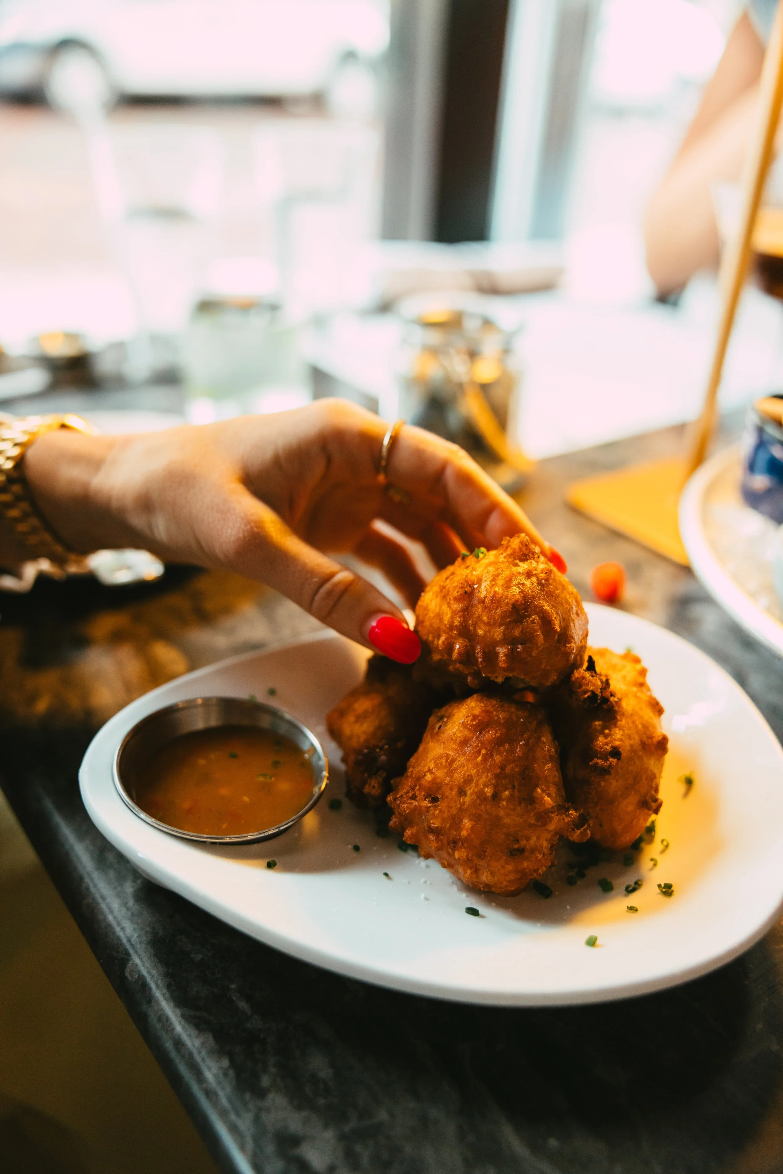 A person with red nail polish and a gold watch is holding a piece of fried food over a white plate with three more fried foods and a small container of dipping sauce.