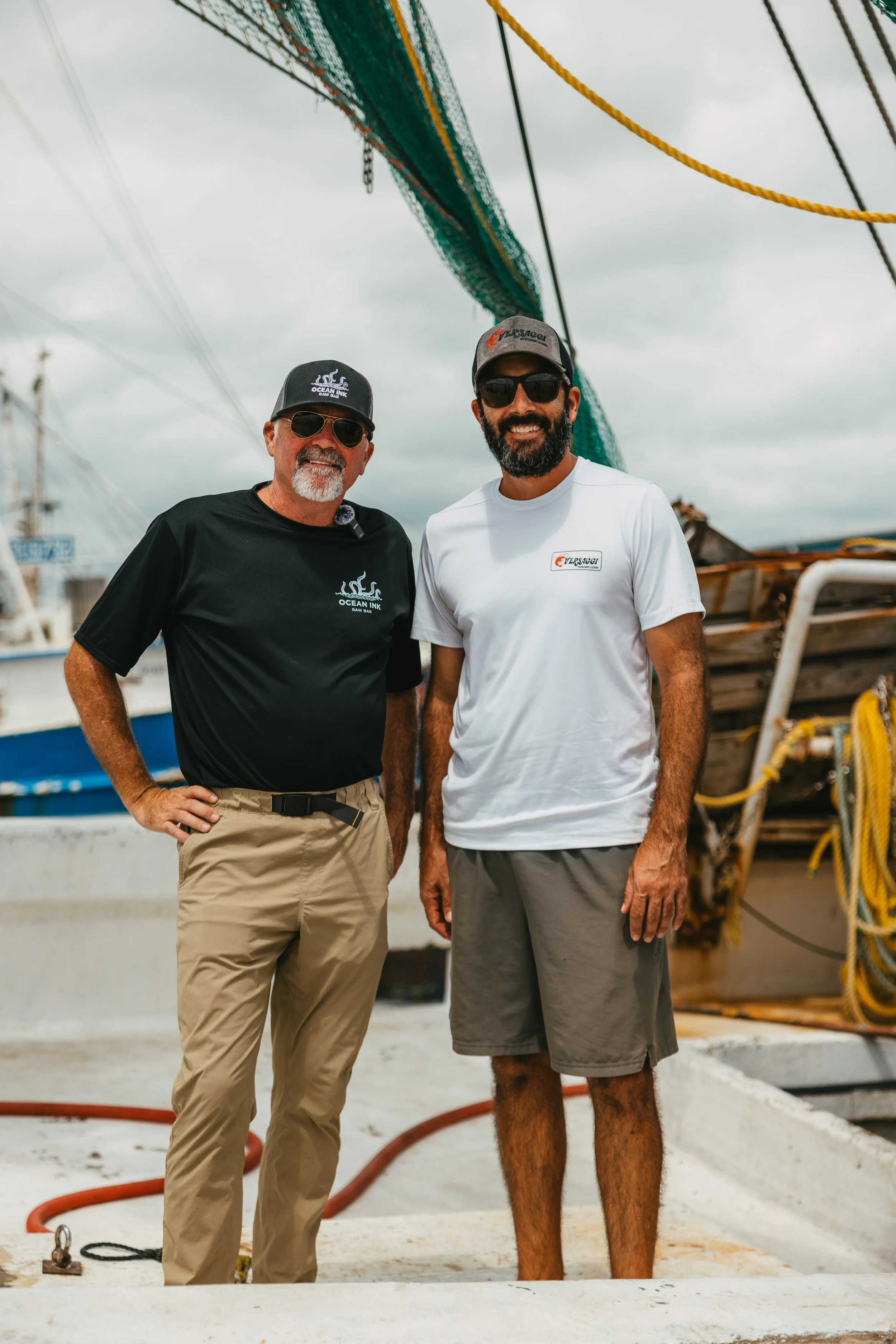 Two men standing on a boat dock with boats in the background. One man wears a black Ocean Ink shirt and khaki pants, the other wears a white shirt and gray shorts. Both are wearing sunglasses and hats, smiling at the camera.
