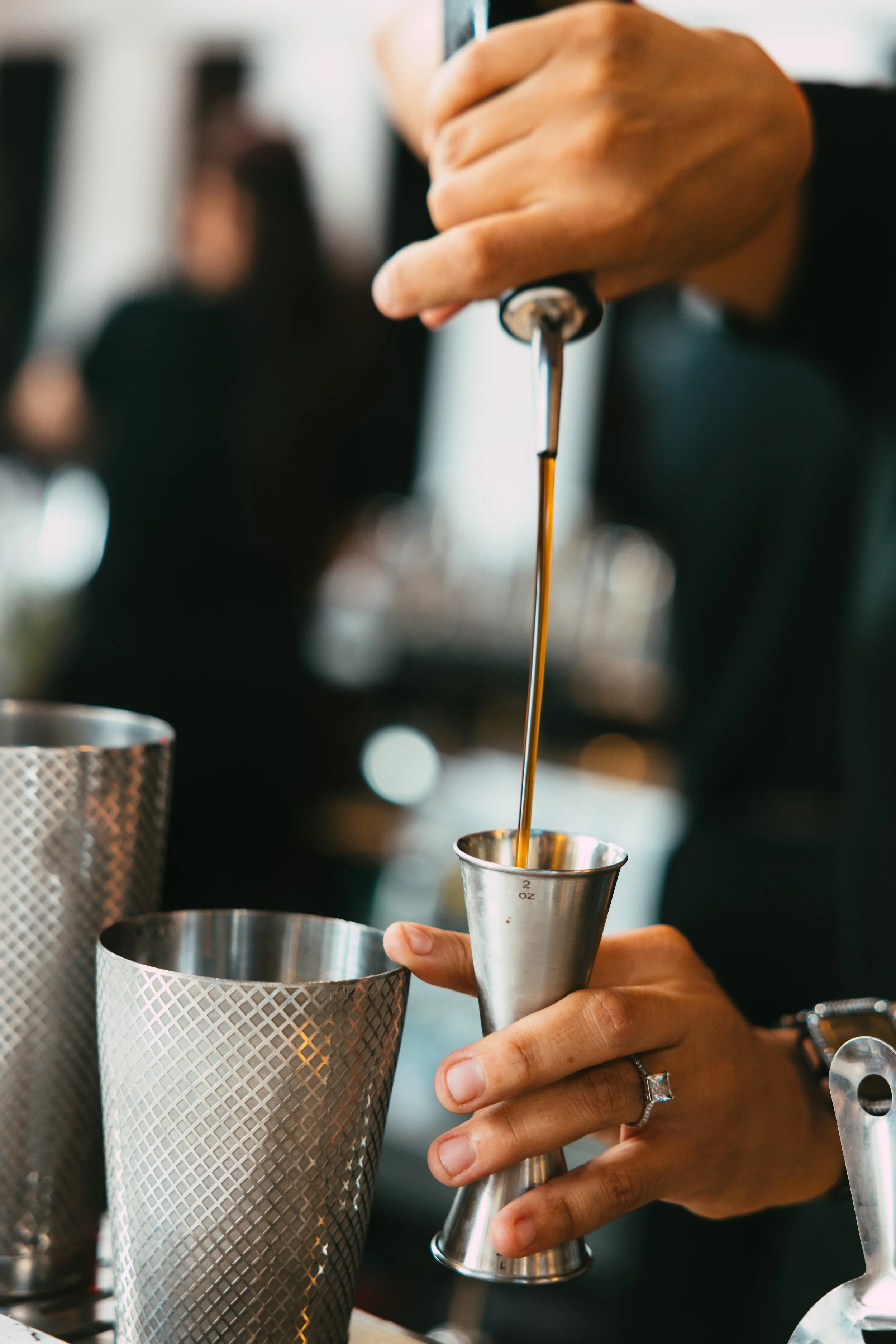 Close-up of a person's hand pouring a dark liquid, likely coffee syrup, from a metal dropper into a stainless steel jigger, with other bar tools visible on the bar counter.