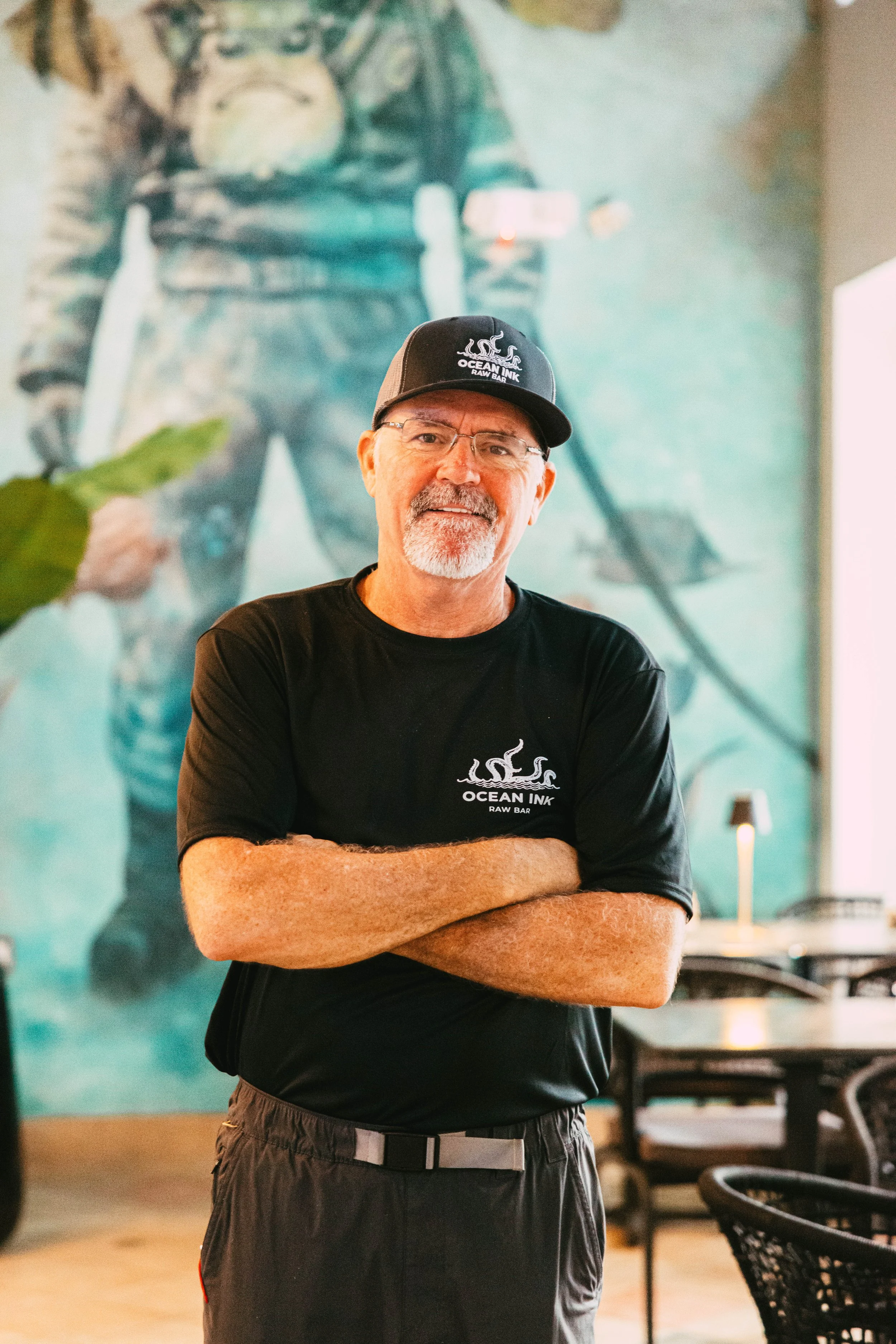 A man with glasses, a beard, and a mustache standing with arms crossed in front of a mural of a person wearing tactical gear. He is wearing a black Ocean Ink T-shirt and a matching cap. The setting appears to be indoors, possibly a restaurant or cafe