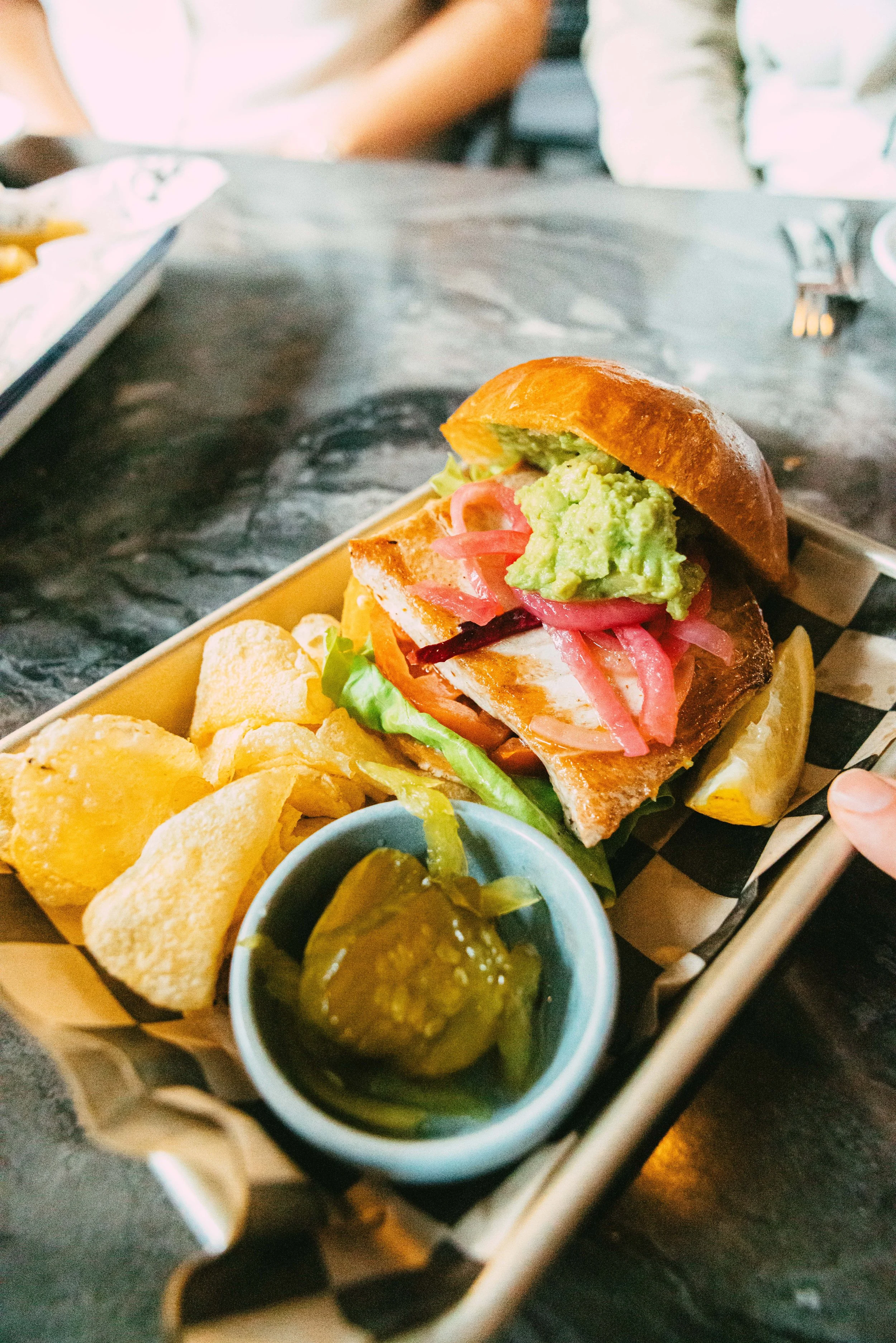 Fish sandwich with guacamole, pickles, potato chips, and pickle slices on a checkered tray.