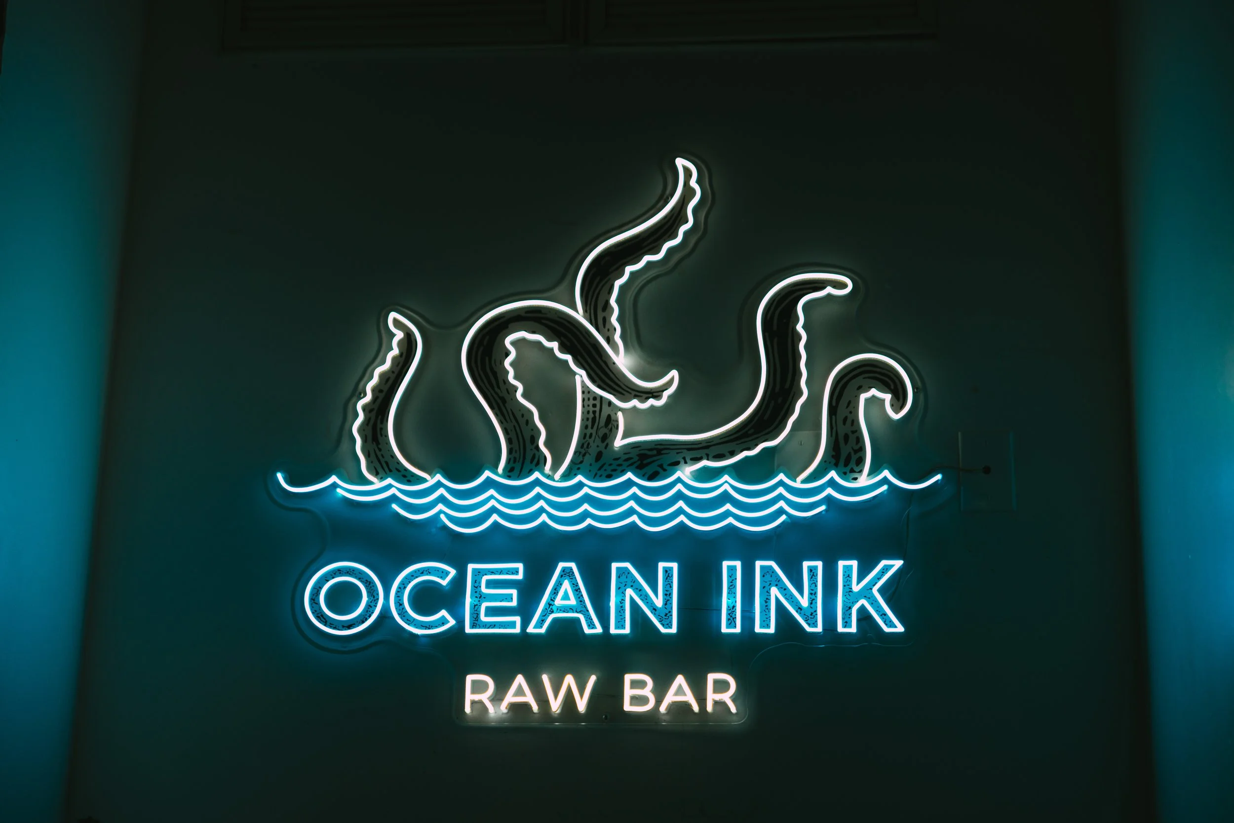 Neon sign of octopus tentacles above the text "OCEAN INK RAW BAR" on a dark wall.