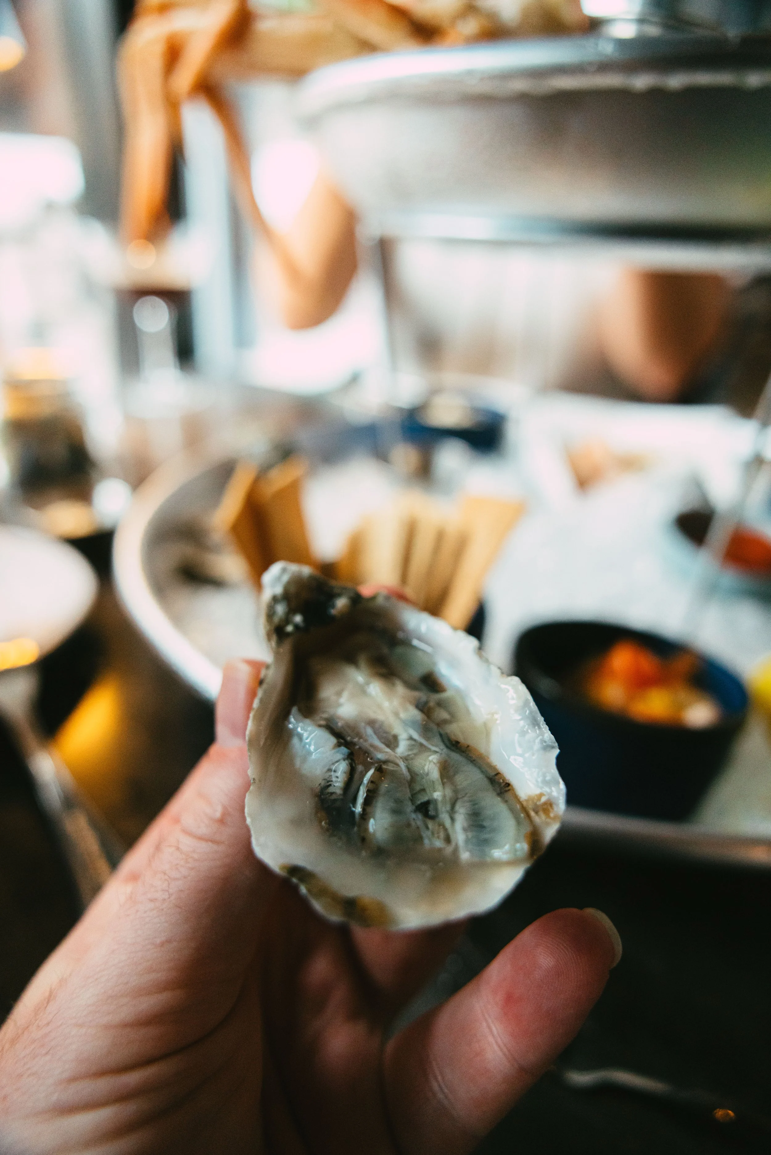 Person holding a raw oyster shell over a seafood buffet with various dishes and utensils visible in the background.
