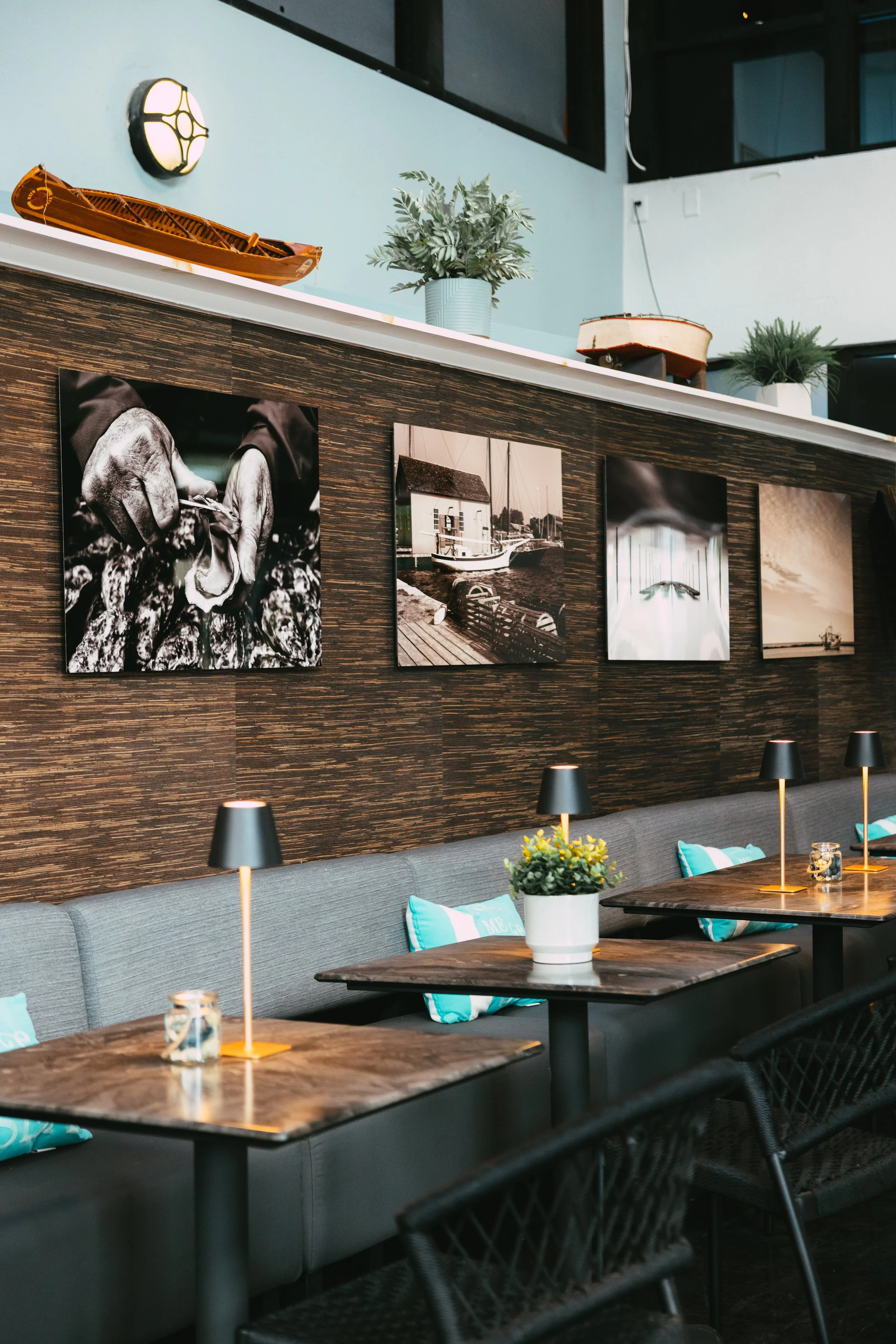 Interior of a cafe or restaurant with wooden tables, black lamps, gray seating, and framed black-and-white photos on a textured brown wall, decorated with potted plants.