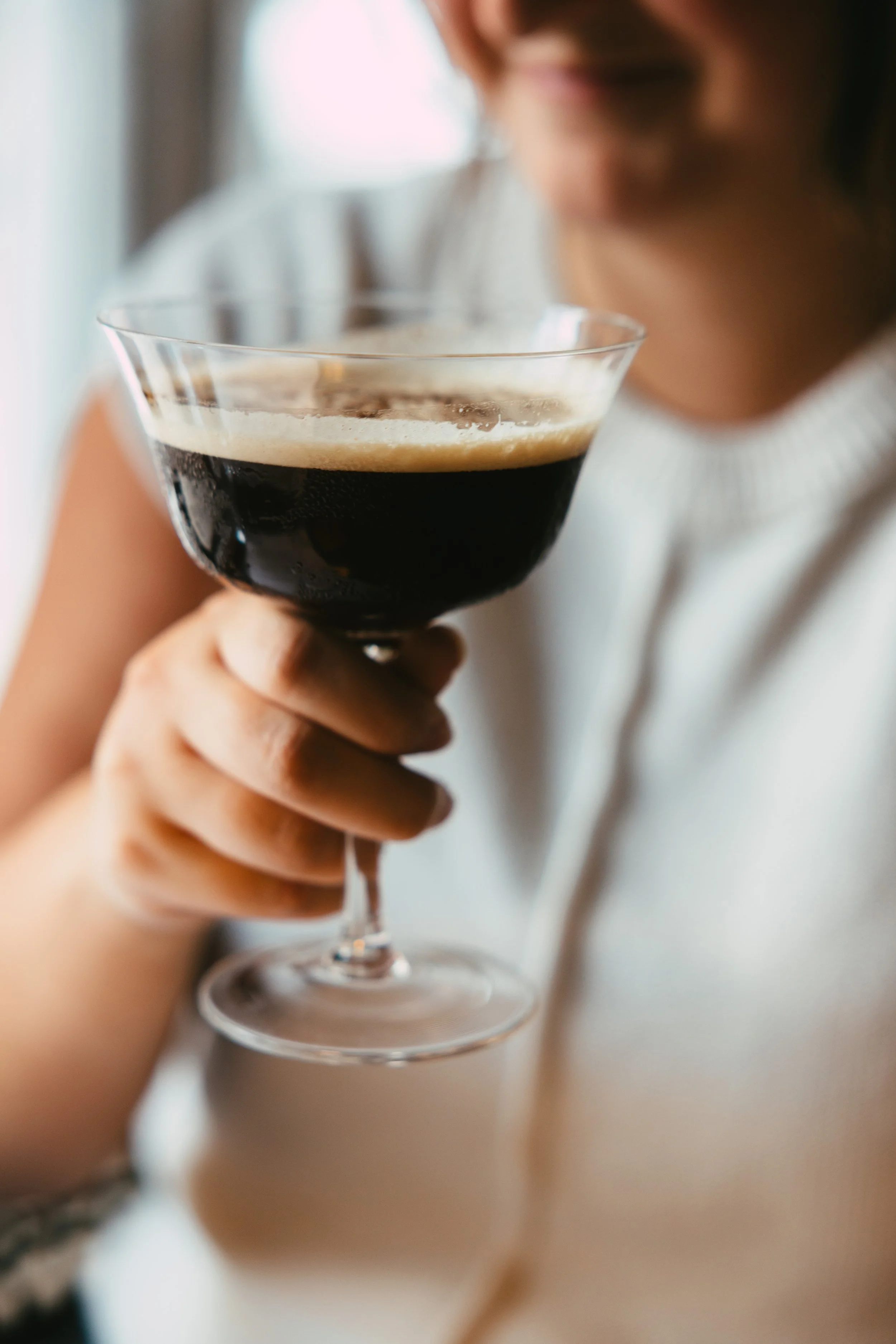 A person holding a glass of dark coffee or espresso with a layer of crema on top.