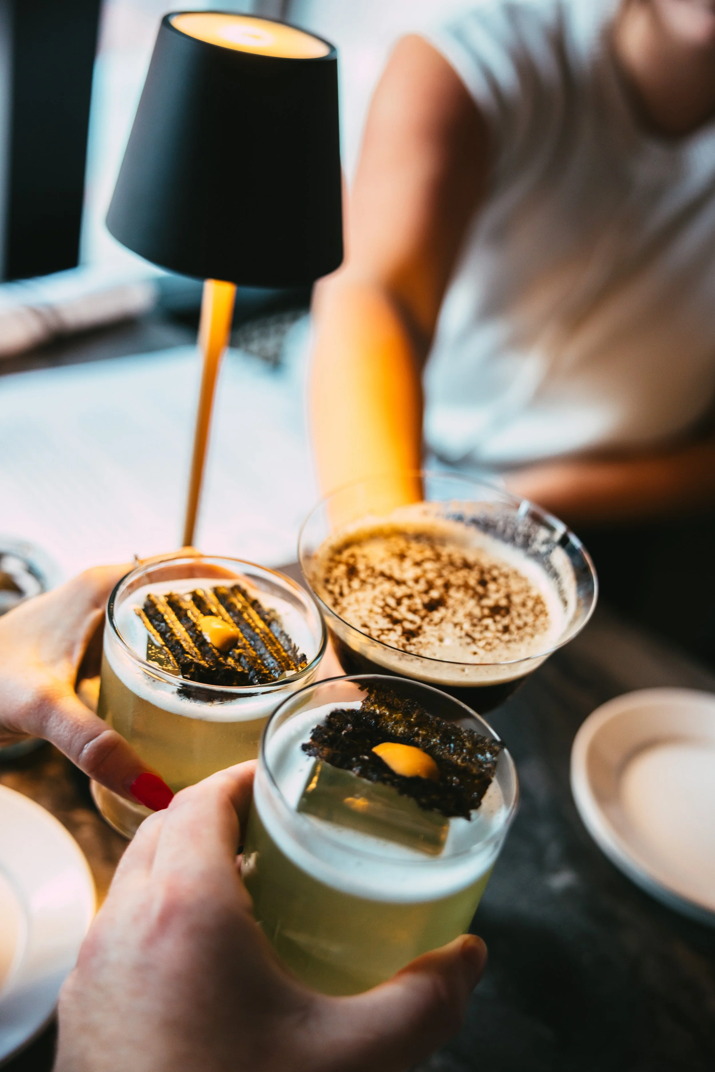 Two people hold glasses with a layered green cocktail topped with a black seaweed cracker and a dollop of yellow sauce, with a person in the background pouring drinks, and a table with extra glasses and a lamp.