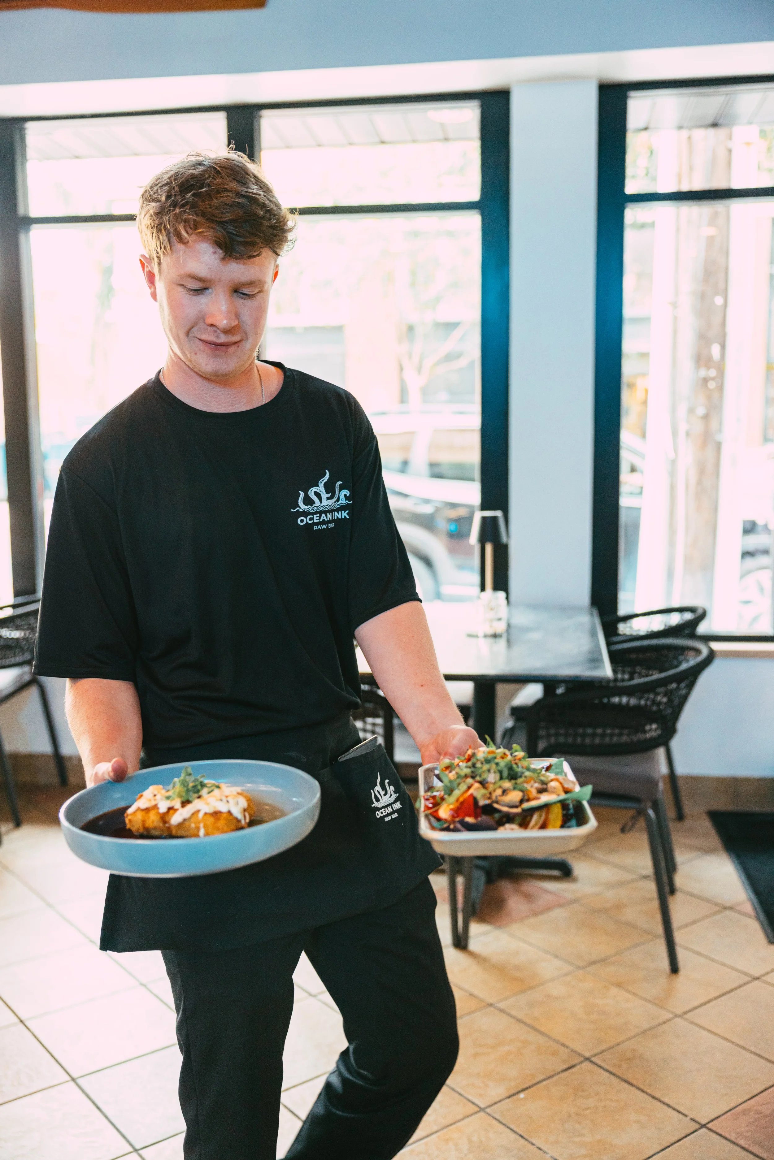 A young man in black uniform holding two plates of food, inside a well-lit restaurant with large windows, black chairs, and tiled floor.