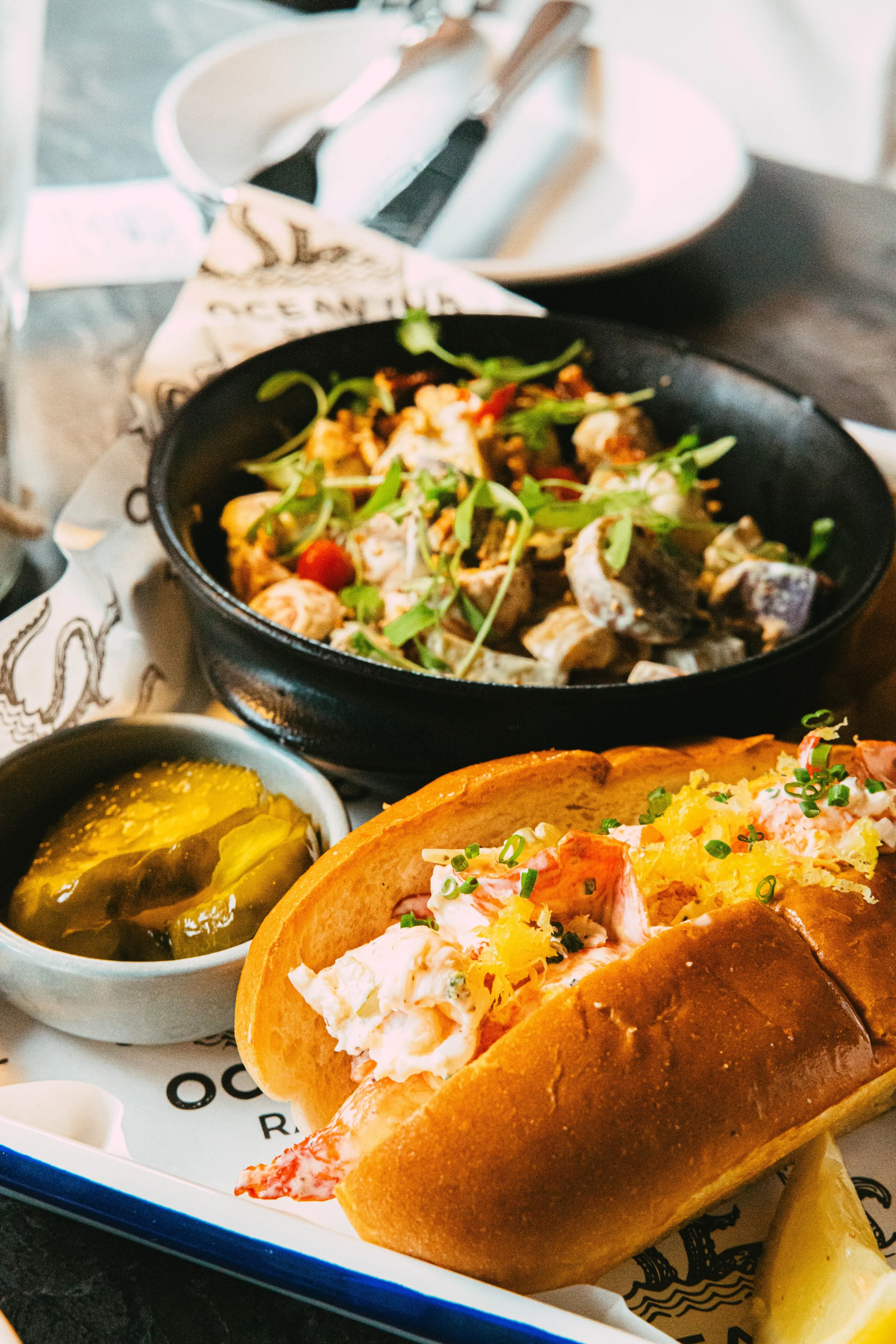 Lobster roll in a toasted bun, side of pickles, and a vegetable dish with greens and cherry tomatoes on a tray.