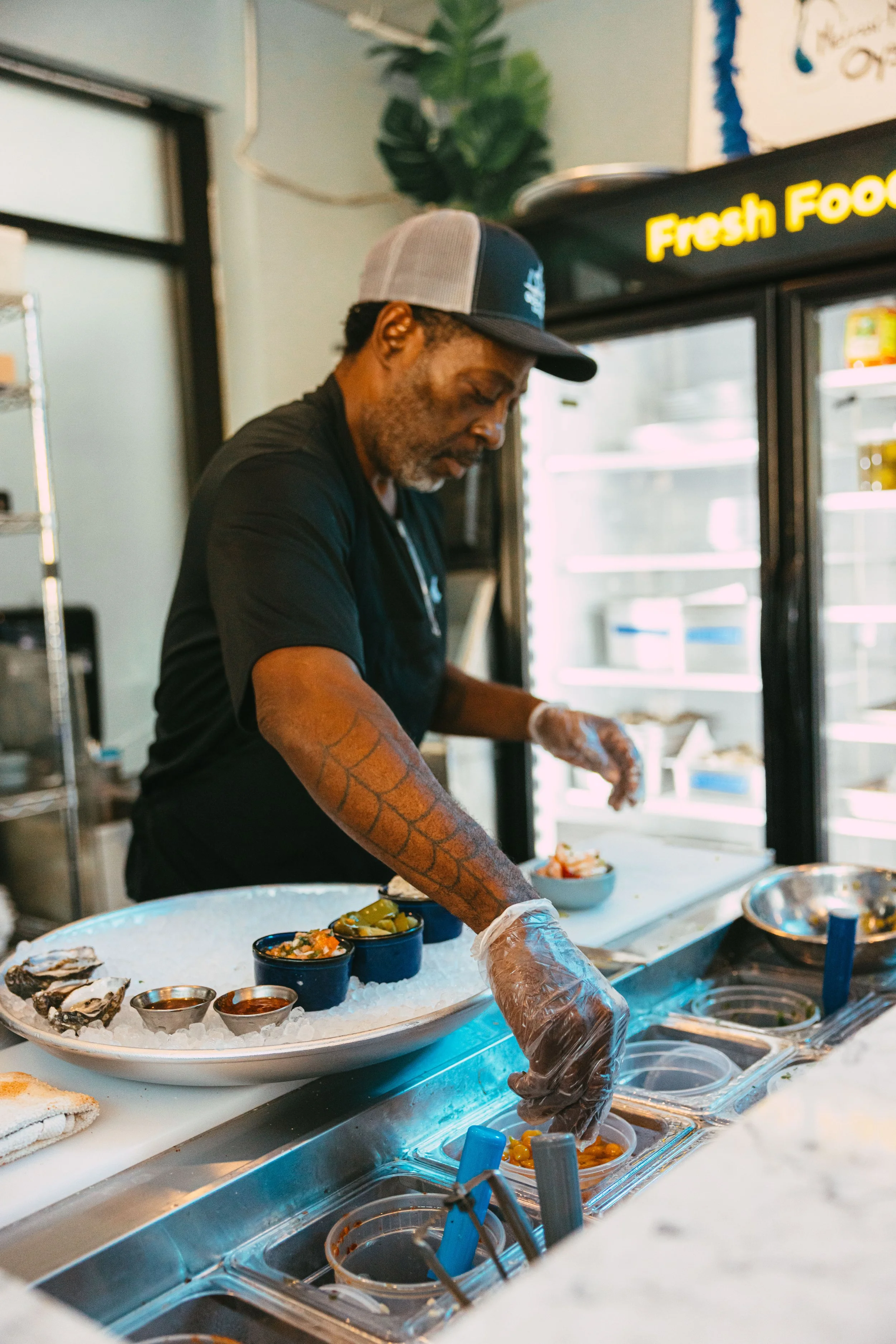 A man wearing a black shirt, a baseball cap, and plastic gloves is preparing food behind a counter with bowls of condiments, seafood, and other ingredients. There is a refrigerator with a stick-on sign that says 'Fresh Food' in the background.
