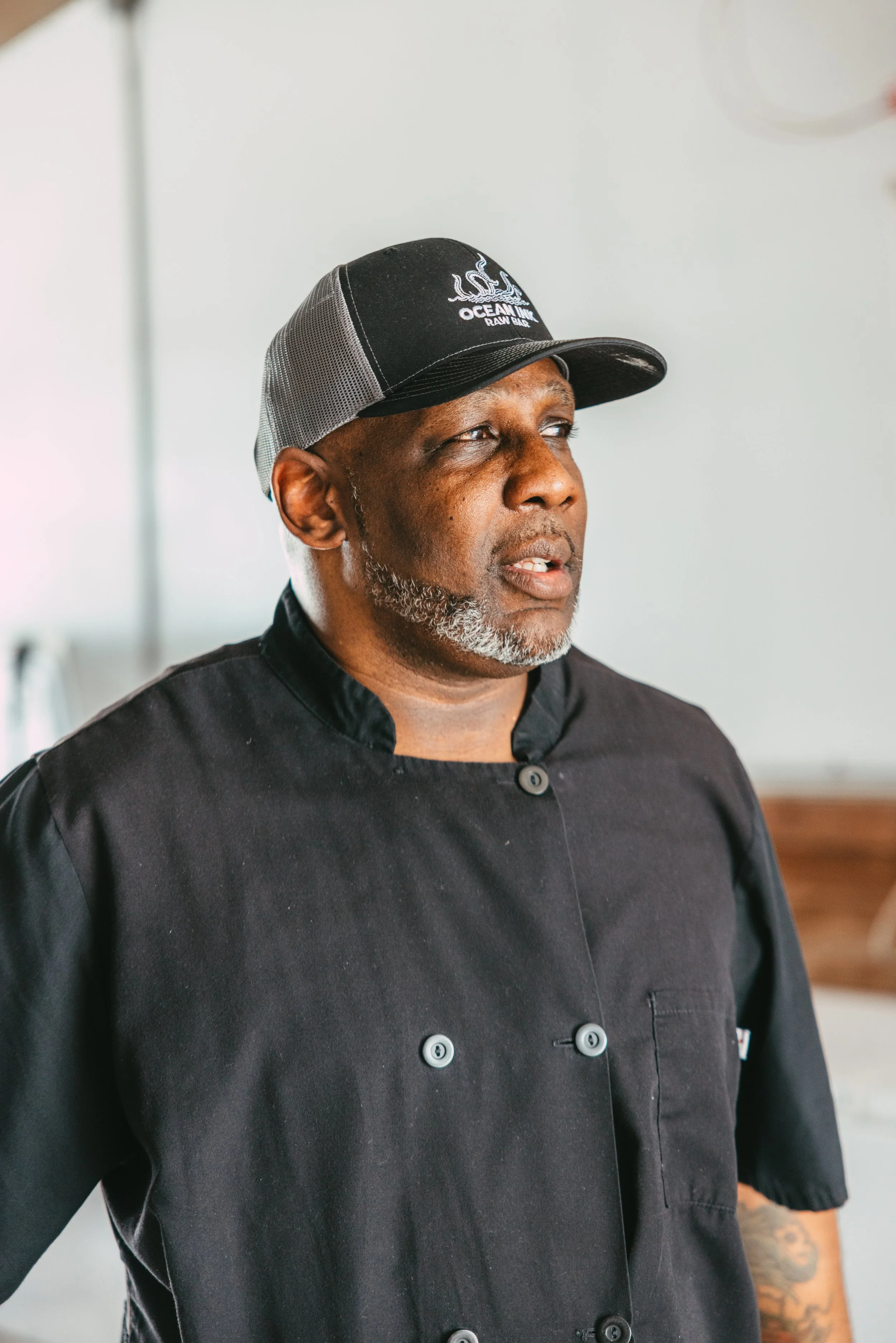 A middle-aged man wearing a black chef's coat and a baseball cap that says 'OCEAN ISLAND RAW BAR'. He has a gray beard and tattoos on his arm, and is looking off to the side.