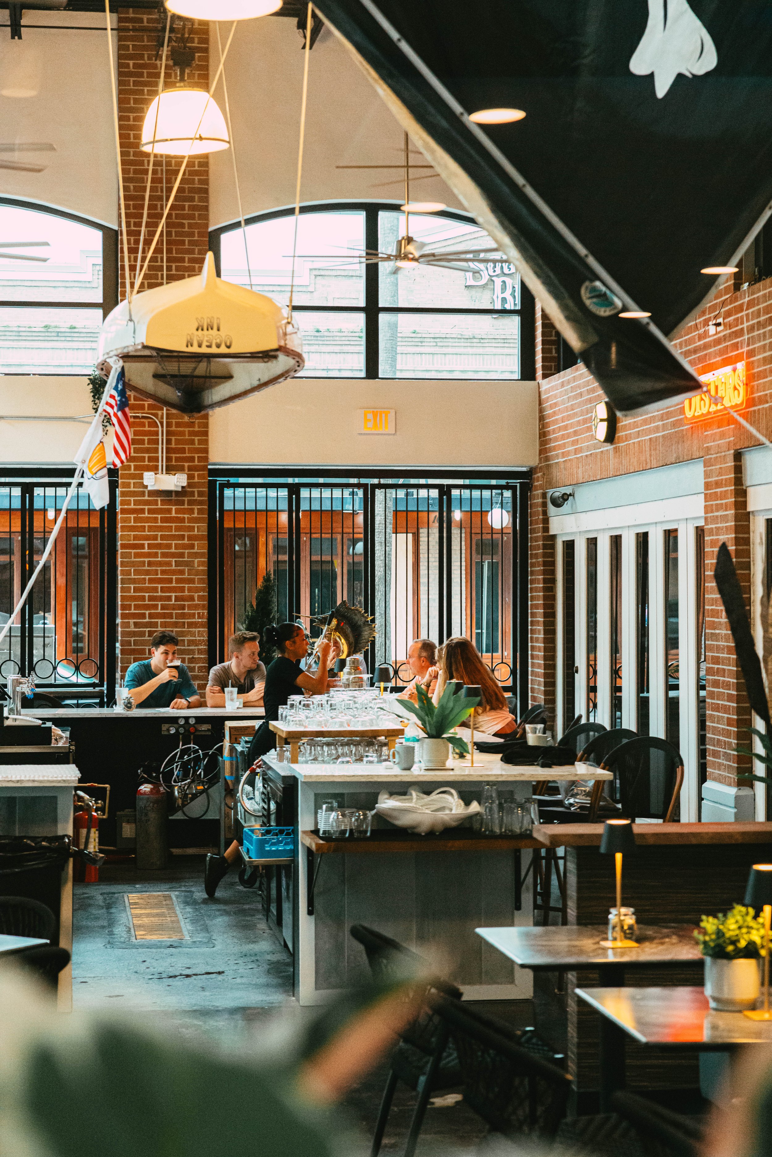 Interior of a restaurant with a group of people sitting at tables and a bar in the background. The decor features brick walls, large windows, and nautical-themed elements, including a boat hanging from the ceiling.