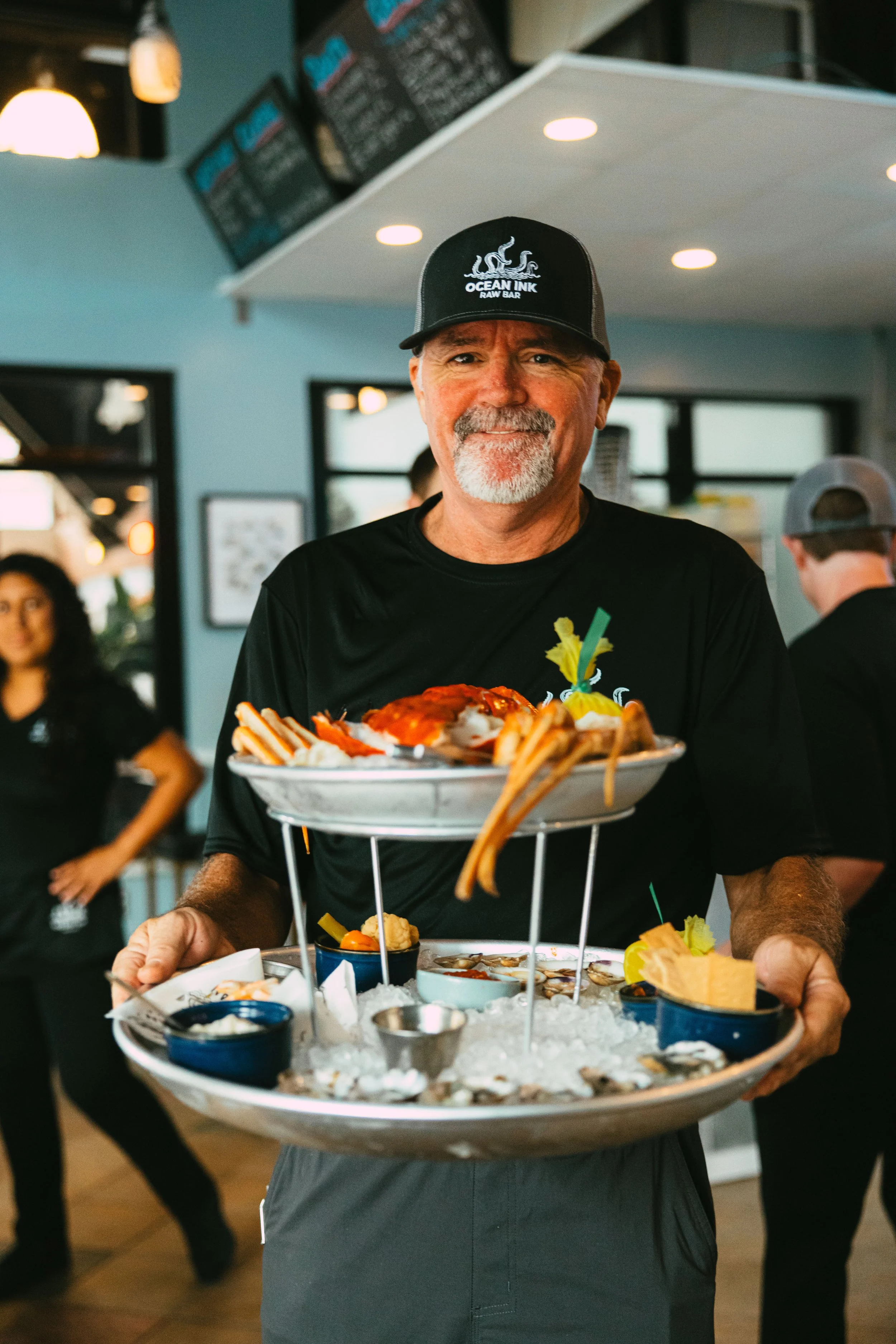 A man wearing a black Ocean Ink hat and black shirt is serving a seafood platter at a restaurant. The platter contains seafood, fries, and lemon wedges, and is placed on a bed of ice. The restaurant has a casual atmosphere with menu boards visible in
