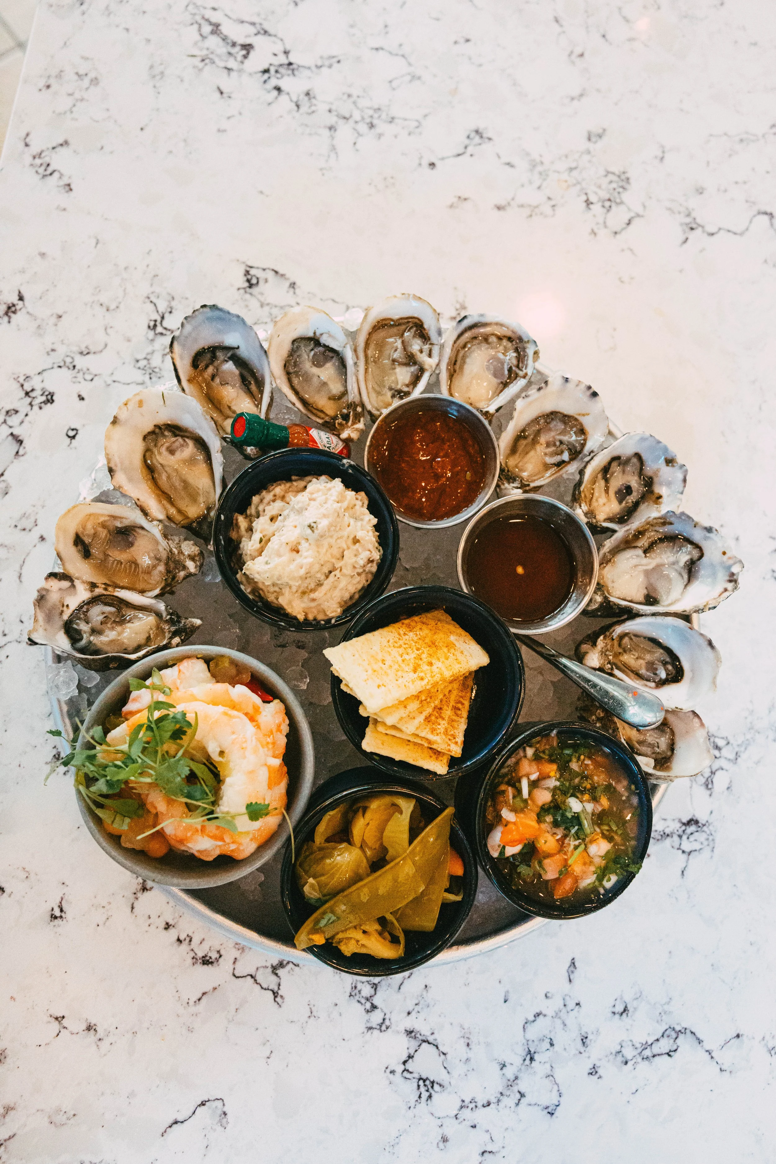 A seafood platter with oysters, dipping sauces, crackers, shrimp, pickles, and a martini glass on a white marble table.