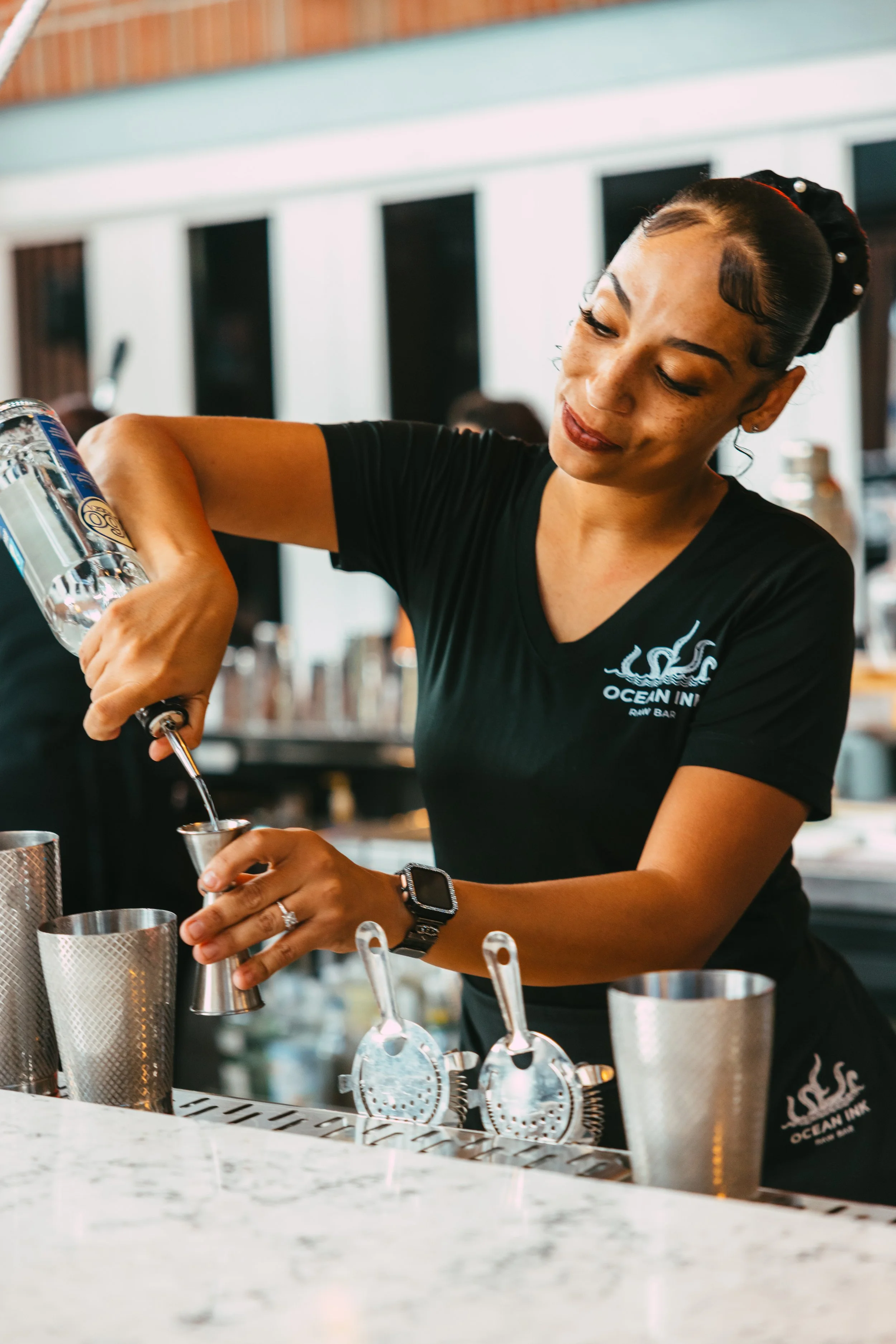 A woman bartender with dark hair in a bun, wearing a black T-shirt with a logo, pouring a clear liquid into a metal jigger at a bar counter.
