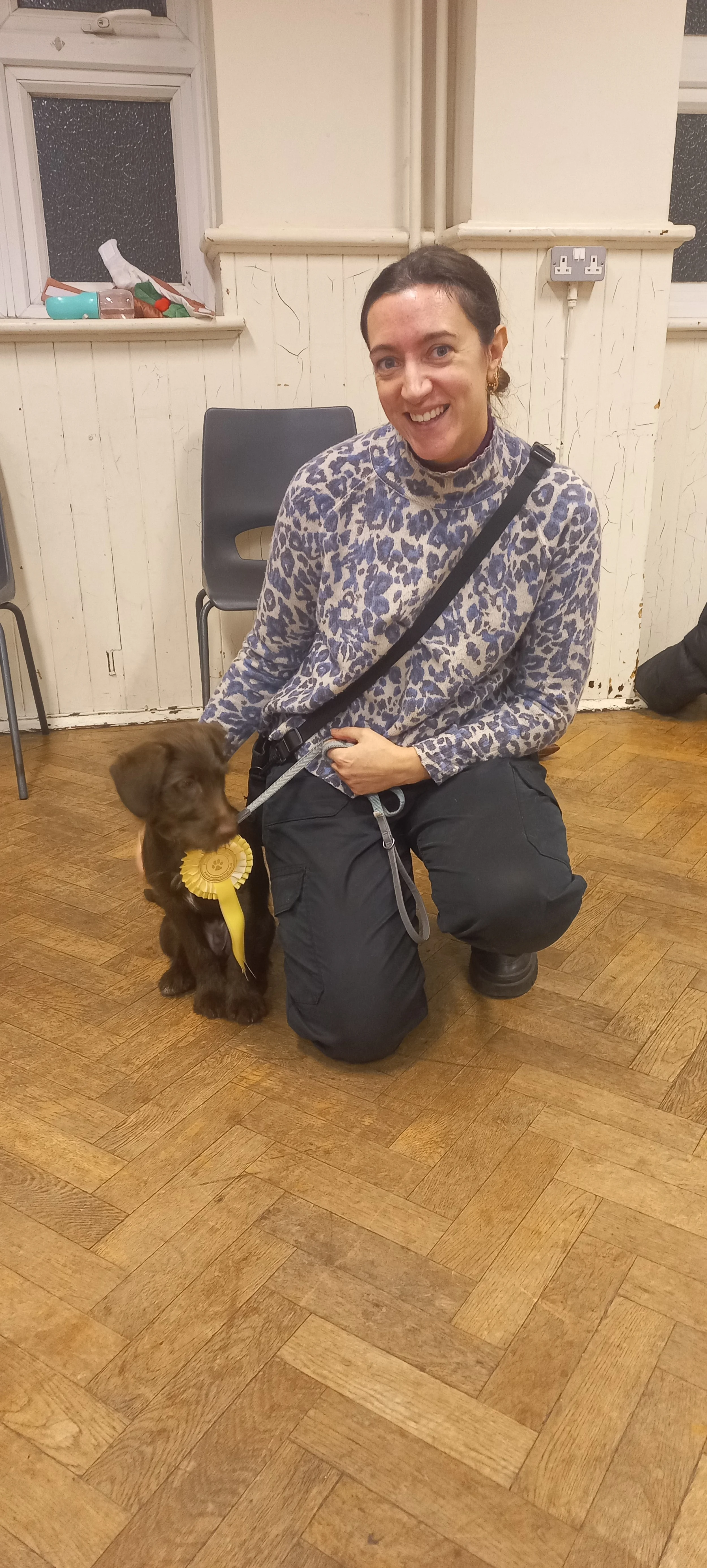 A smiling woman with dark hair kneels on a wooden floor next to a brown puppy with a yellow rosette. The woman wears a leopard print sweater and black pants, and is holding the leash for the puppy.