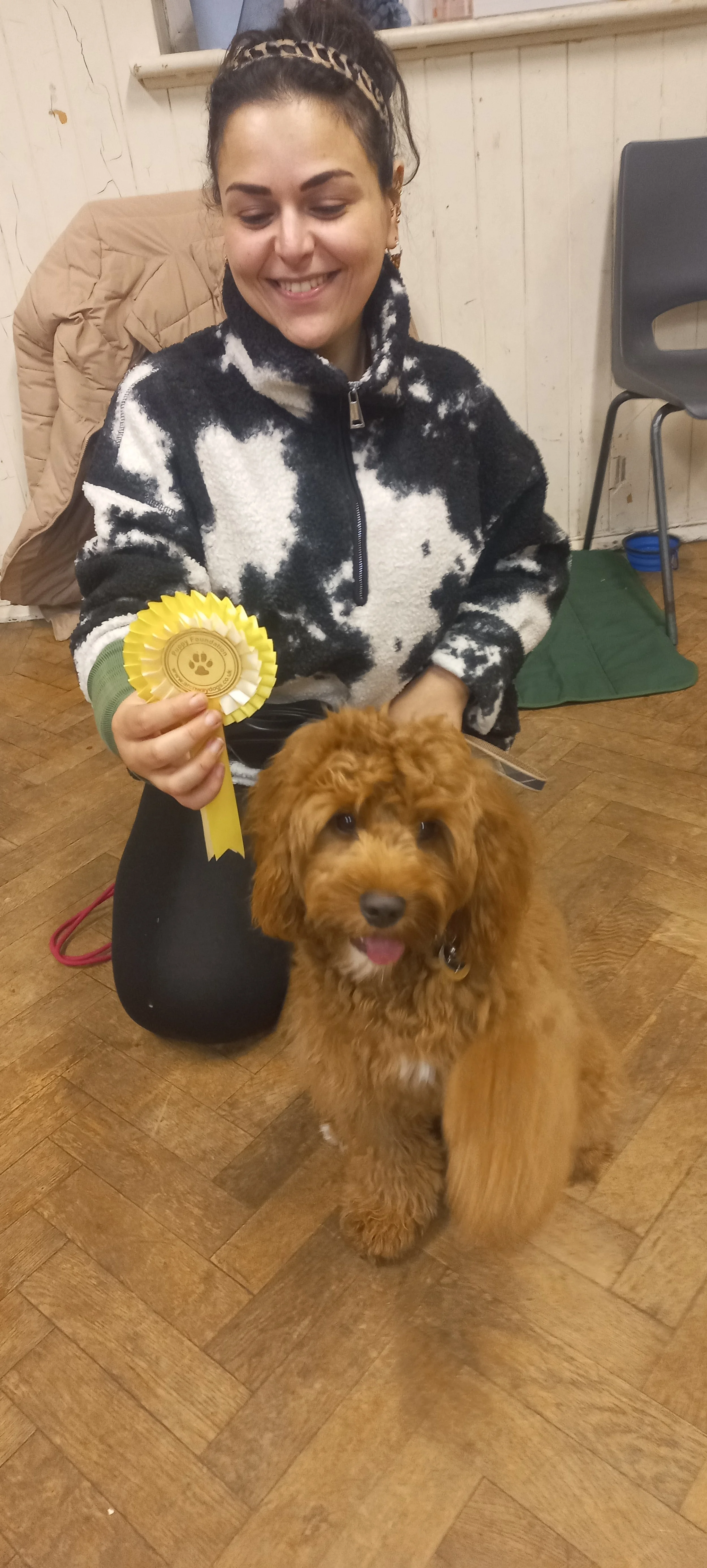 A woman with dark curly hair, wearing a black and white  jacket, is kneeling and holding a yellow rosette with a paw print emblem. A small, fluffy brown dog with curly fur is sitting in front of her, looking at the camera with its tongue out.