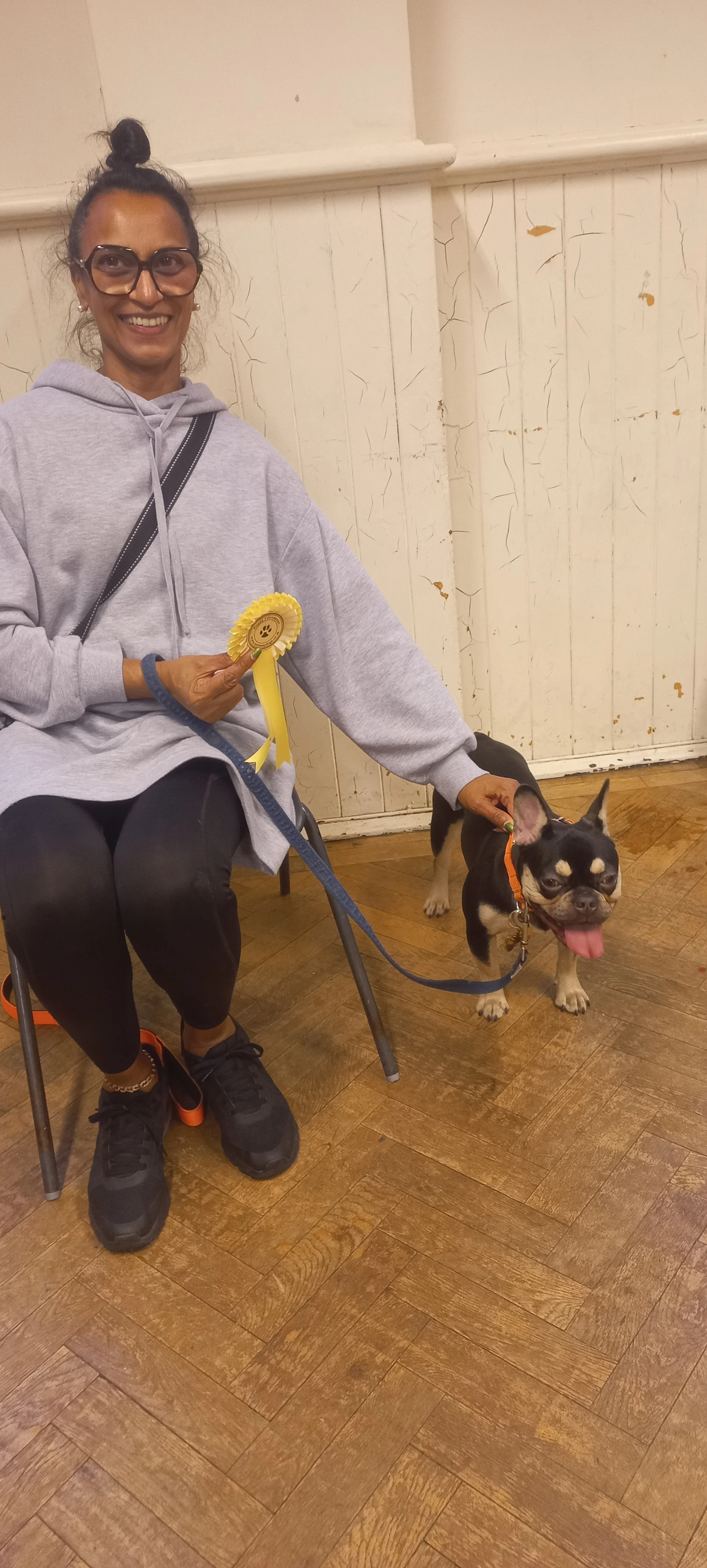 A woman sitting on a chair holding a puppy yellow rosette award, with a small black and tan French Bulldog on a leash, inside a hall with a wooden floor and white walls.