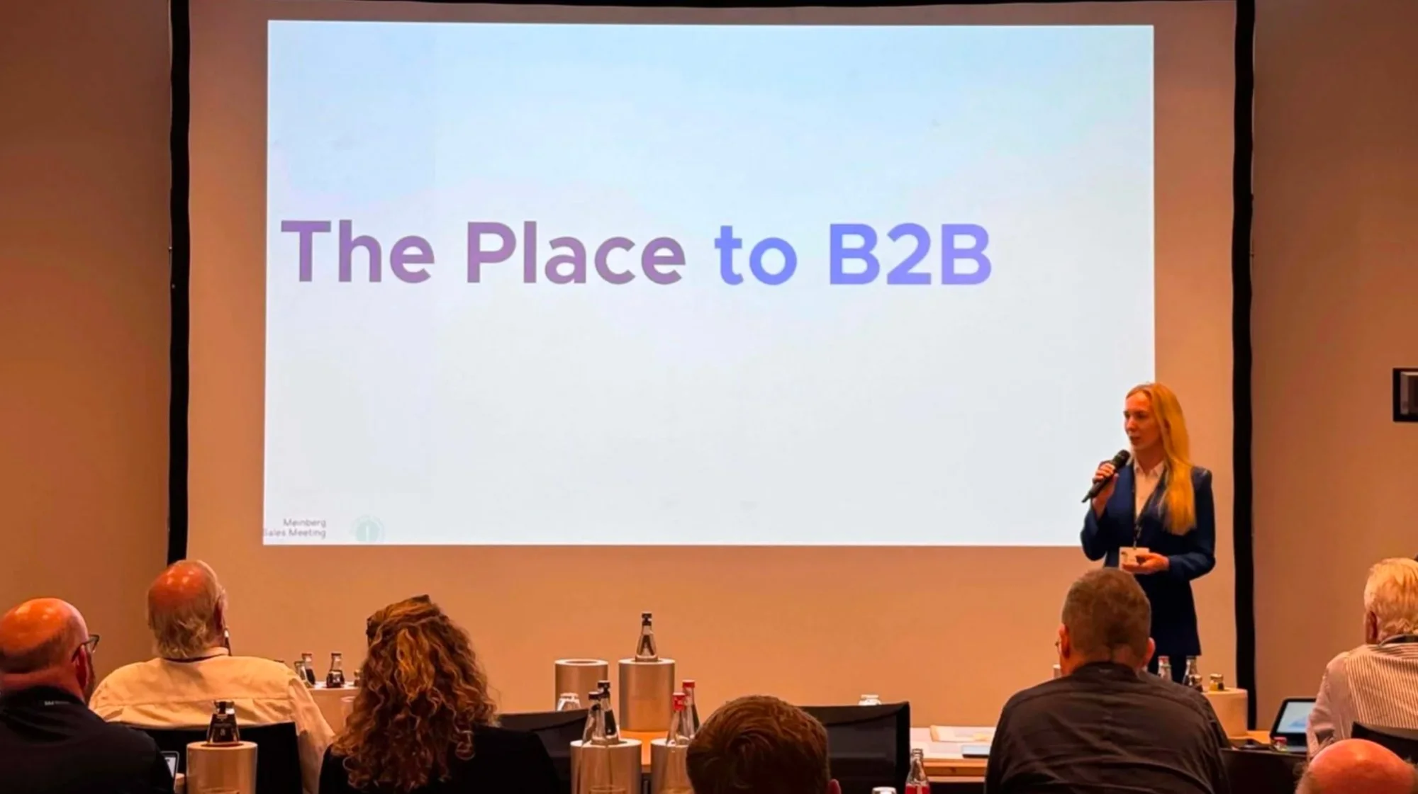 A woman giving a presentation in front of a large screen that reads "The Place to B2B" at a conference or meeting room with attendees seated and taking notes.