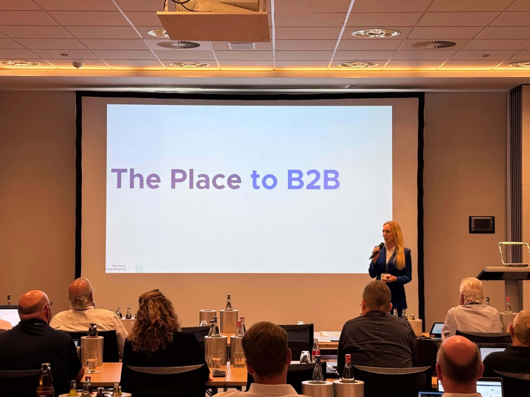 A woman giving a presentation in front of a large screen that reads "The Place to B2B" at a conference or meeting room with attendees seated and taking notes.