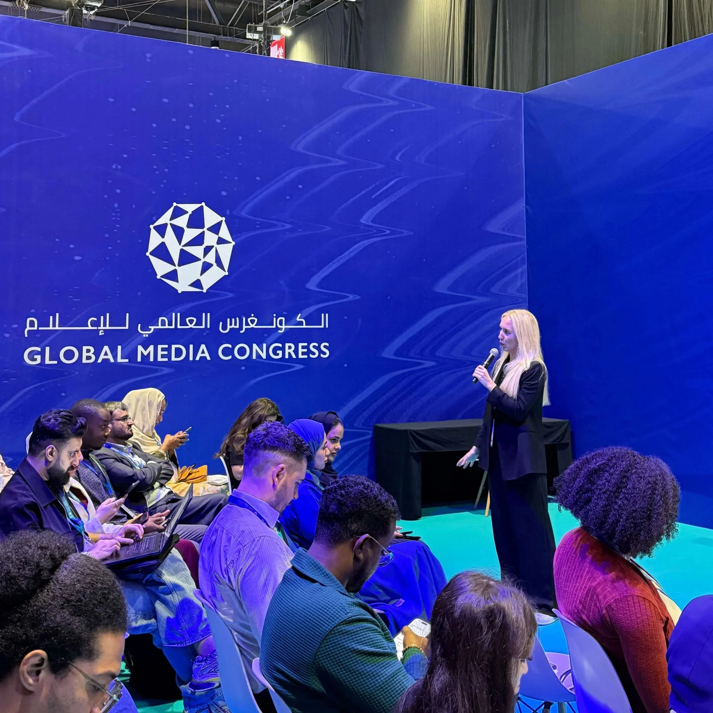 A woman giving a presentation speaking into a microphone at the Global Media Congress, with an audience seated and listening, some taking notes or using laptops, in a conference room with a blue backdrop.