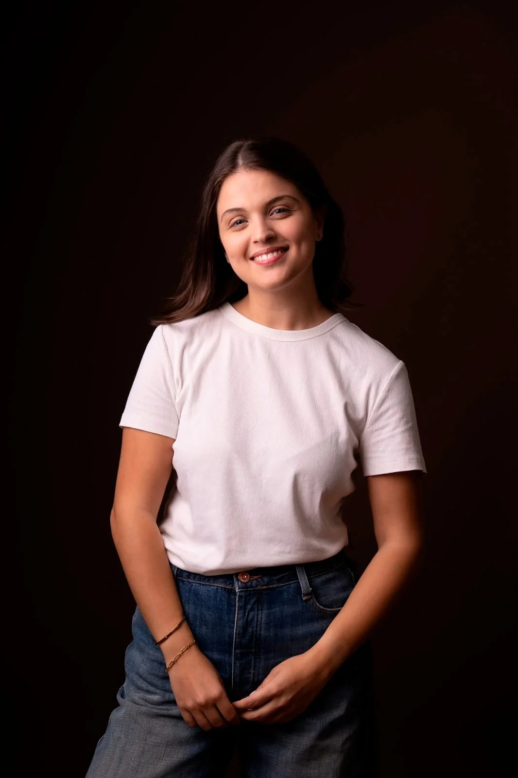 Smiling young woman with dark hair wearing a white t-shirt and jeans against a dark background.