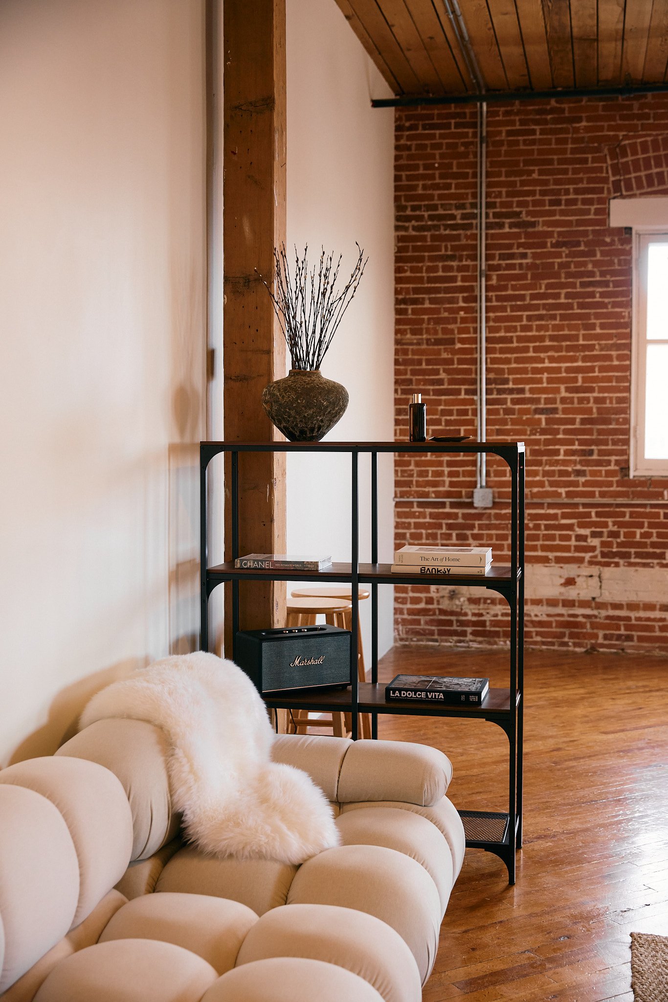A living room with a cream-colored tufted sofa with a fluffy white throw, a black metal bookshelf holding decorative books and objects, a brick wall and window, and a wooden ceiling.