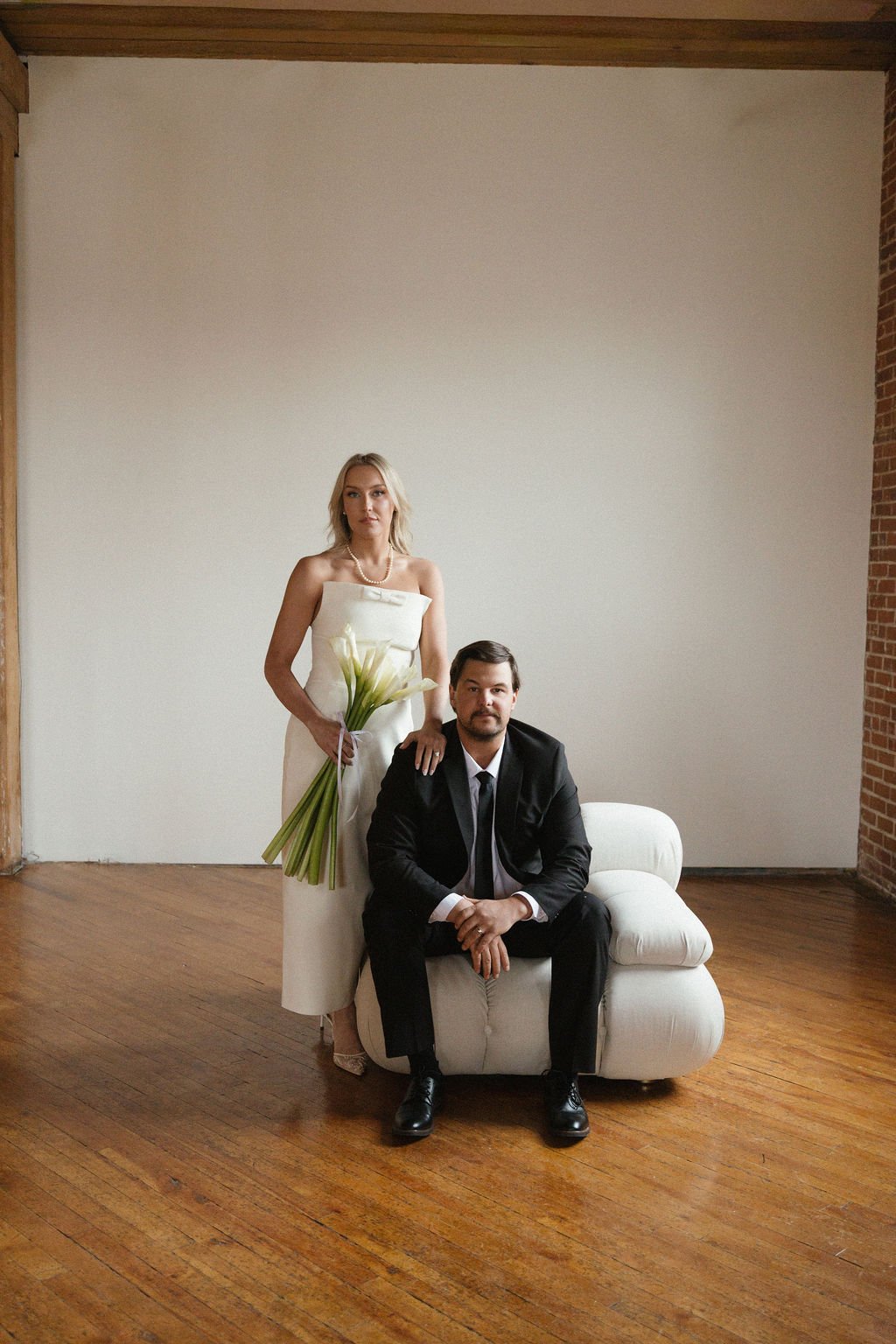 A woman in a white wedding dress holding white calla lilies stands beside a man in a black suit sitting on a white chair in a minimalist room with wooden floors and a large blank wall.