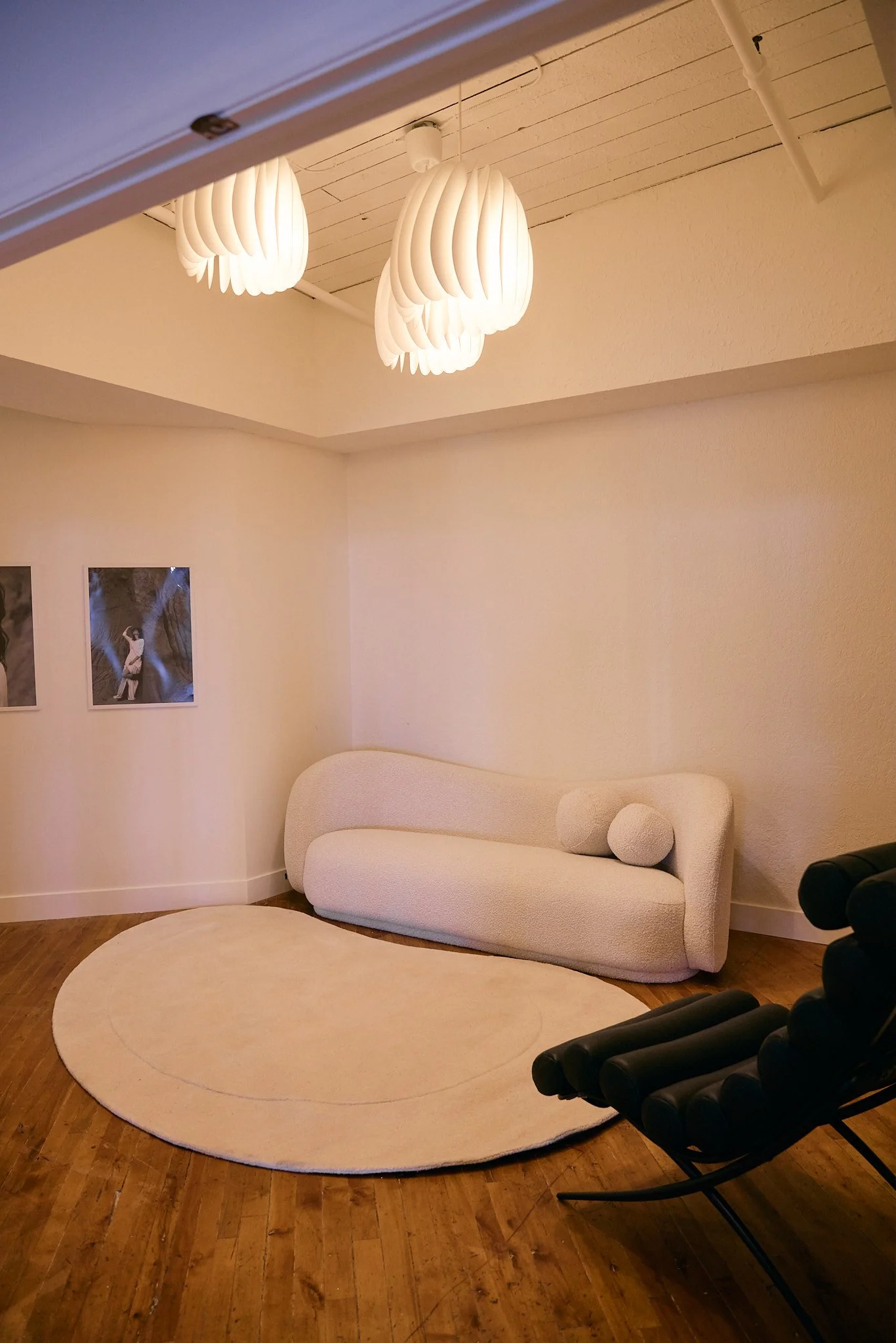 A modern living room with a white couch, a round beige rug, and black lounge chairs. Two wall art photos are visible, and the ceiling has contemporary hanging lights.