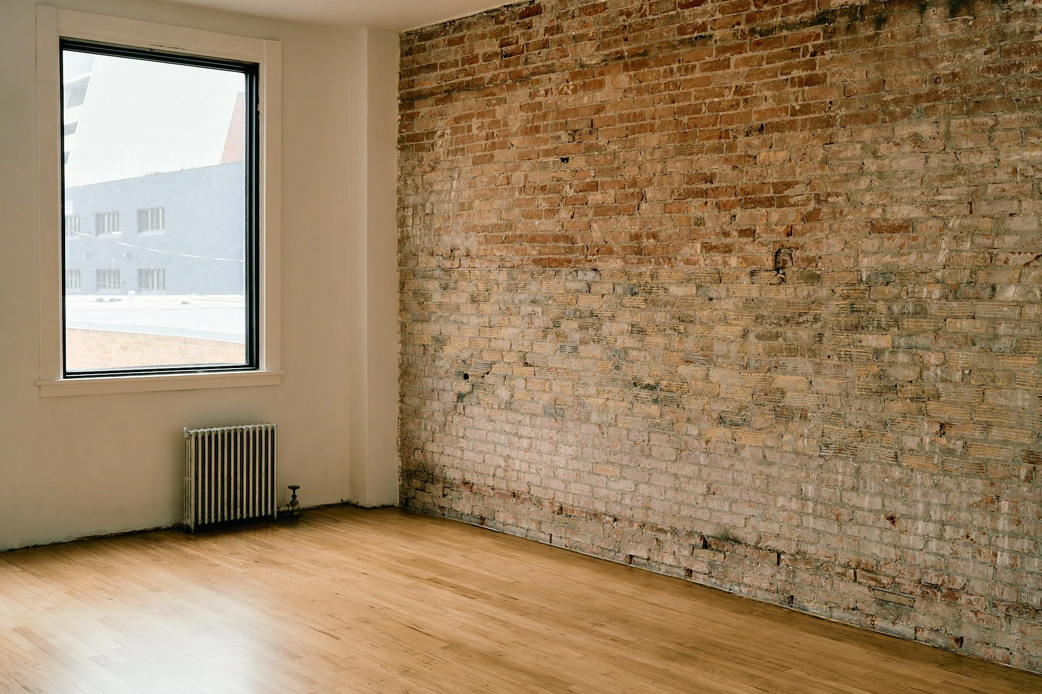 Empty room with a brick accent wall, wooden floor, large window showing neighboring buildings, and a radiator below the window.