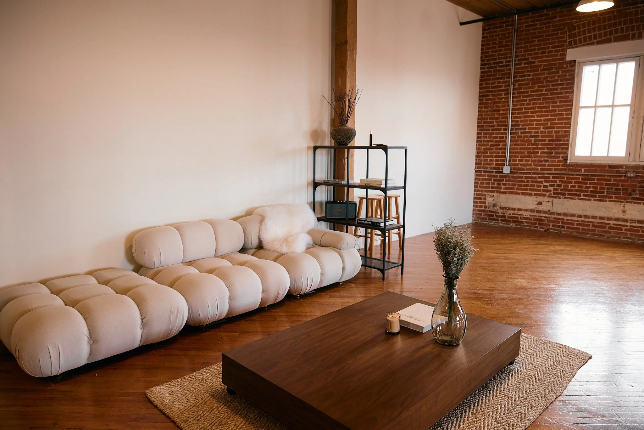 Living room with a beige tufted sofa, a wooden coffee table with a glass vase and a book, a black metal bookshelf with decorative items and books, exposed red brick wall, white wall, window with white frame, hardwood floors, and a beige rug.