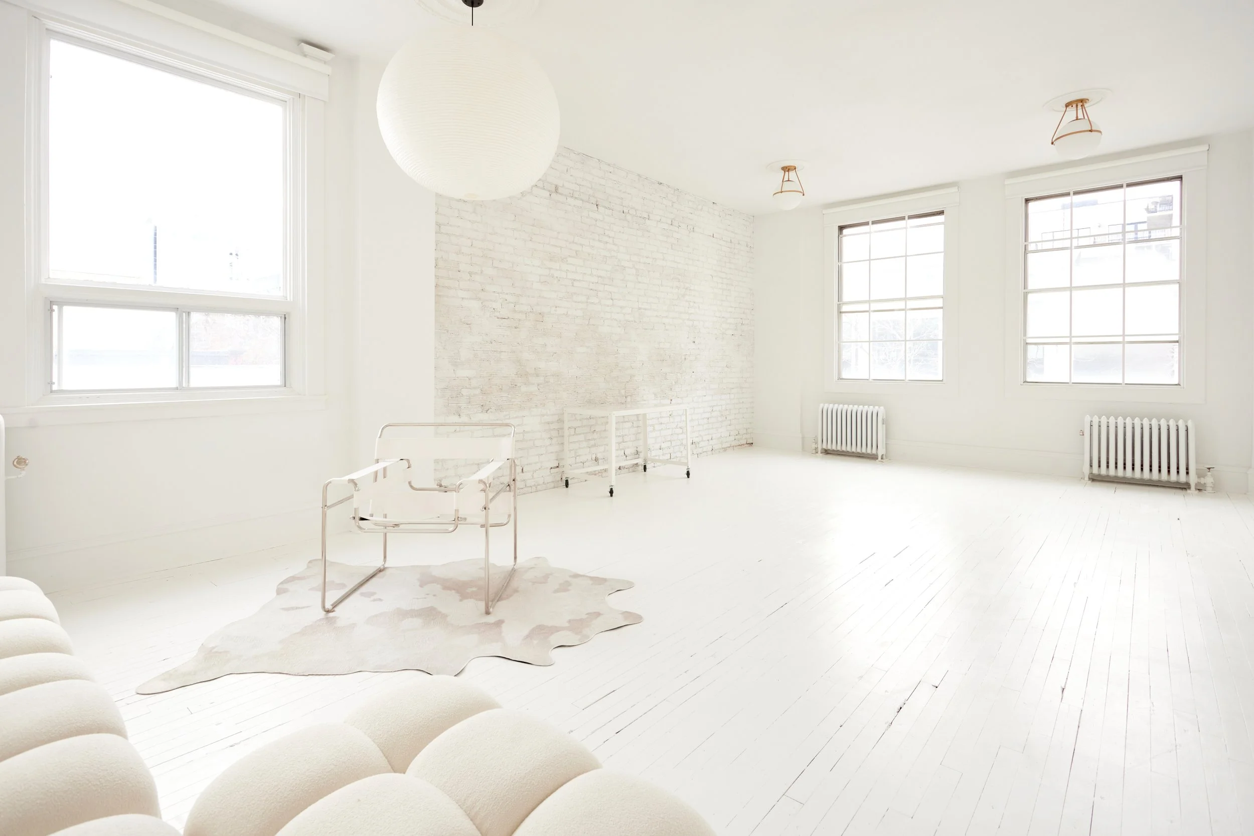 Bright, minimalist living room with white walls, large windows, a white brick accent wall, and light wood floors. Furniture includes a light-colored sofa, transparent chair on a cowhide rug, and white radiators under the windows. Ceiling lights are s