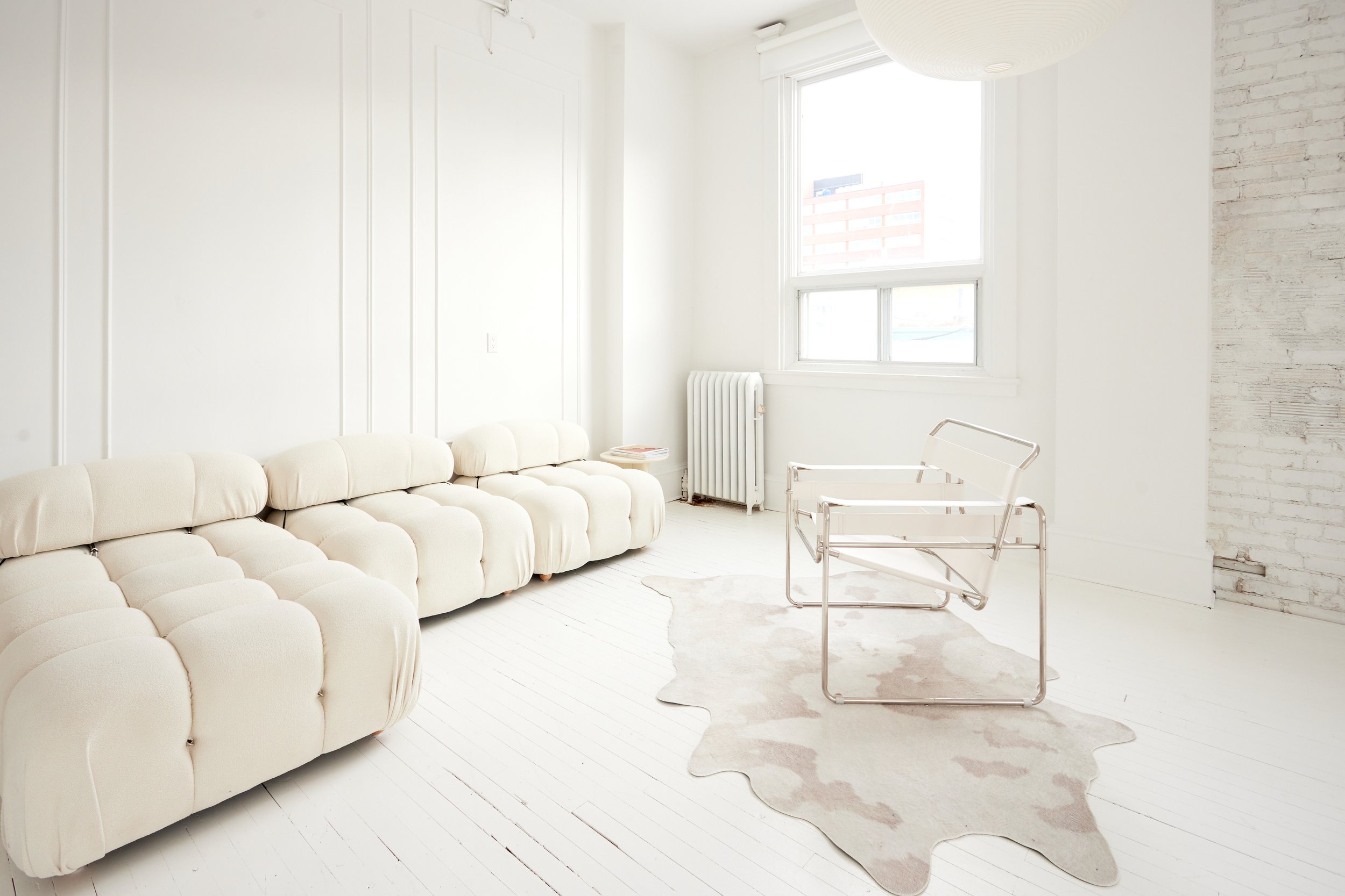 Bright, minimalistic living room with cream-colored sectional sofa, white walls, a window with city view, a modern clear acrylic chair, a light-colored cowhide rug, and a bare white brick wall.