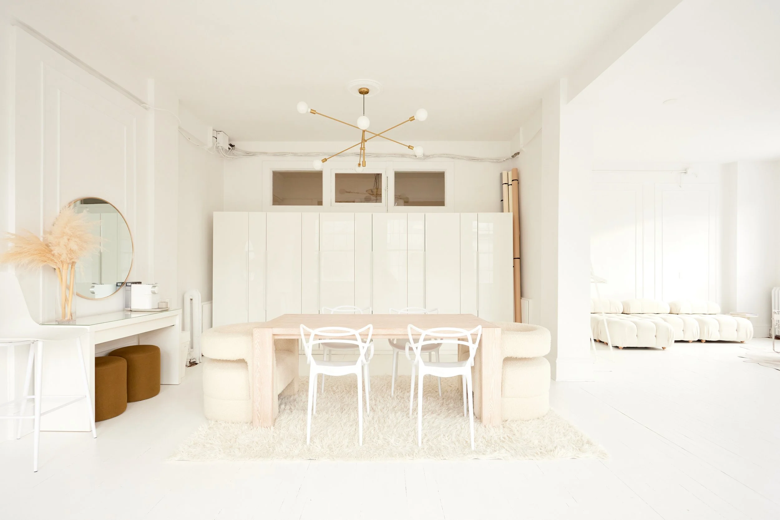 Bright, minimalist white living room with a wooden dining table, white chairs, a cozy beige banquette, and a white tufted sofa. Modern chandelier hangs from the ceiling, and decorative items include a round mirror, a vase with pampas grass, and a cabinet. Large windows and neutral tones create an airy atmosphere.