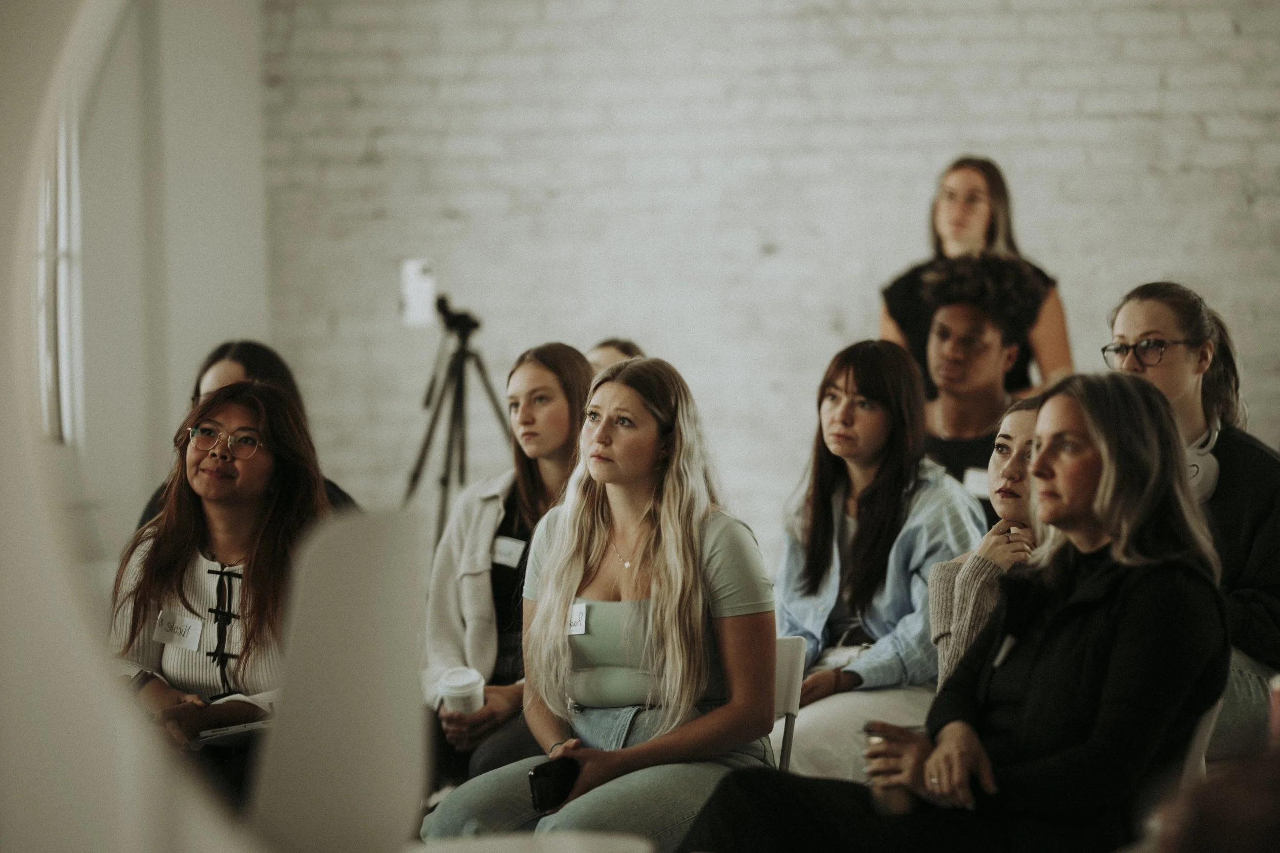 A group of women attending a seminar or conference, sitting and listening attentively, with a woman standing in the background near a tripod.