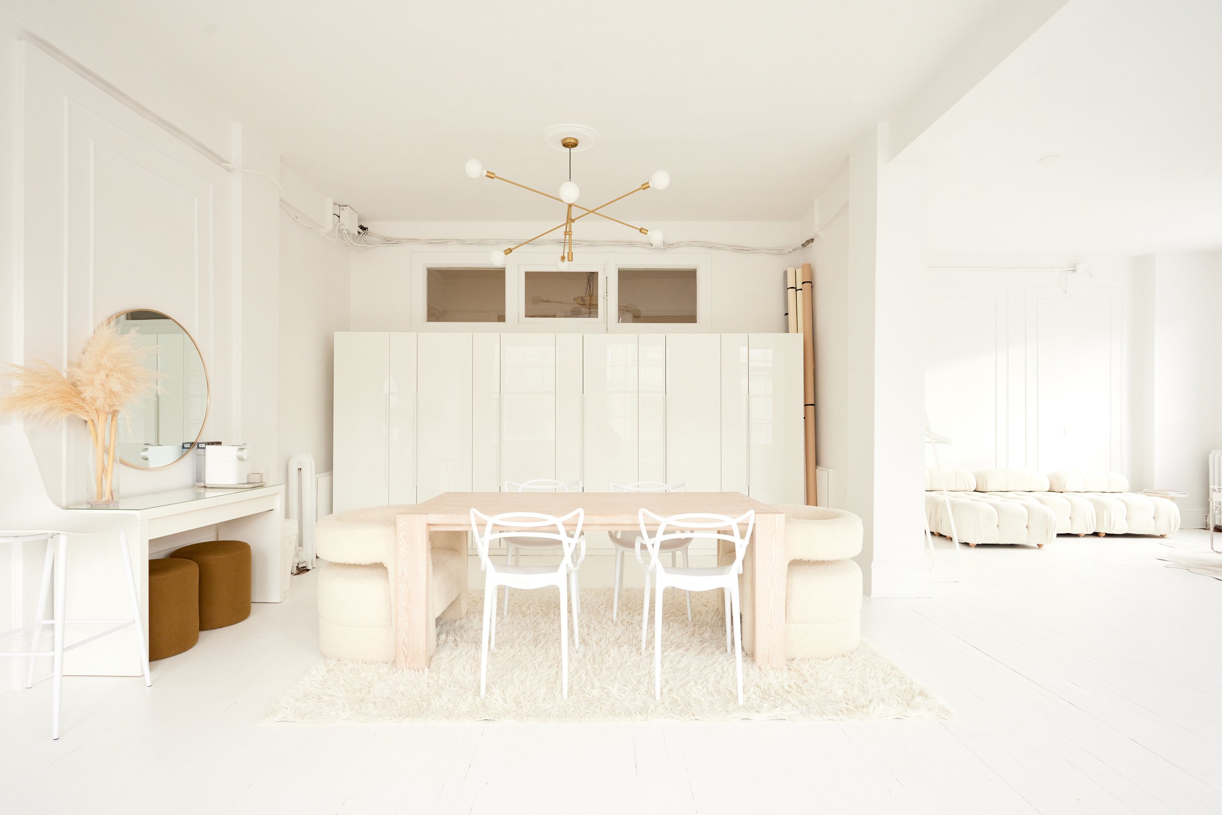 Bright, minimalist living and dining area with white walls, furniture, and decor, a round mirror on left, a dining table with white chairs, and a white sofa in the background.