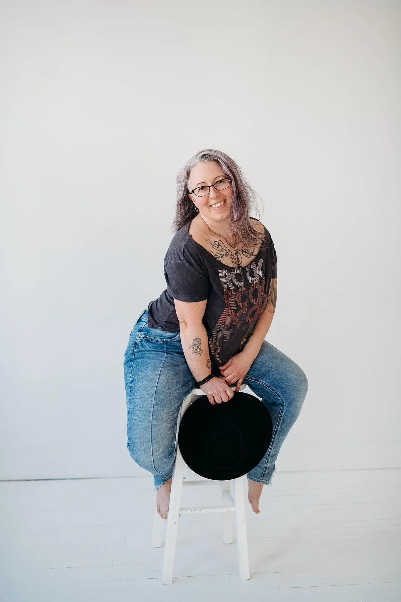 Person sitting on a white stool, wearing a black t-shirt with a design, blue jeans, and holding a black hat. They have tattoos and glasses, and are smiling.