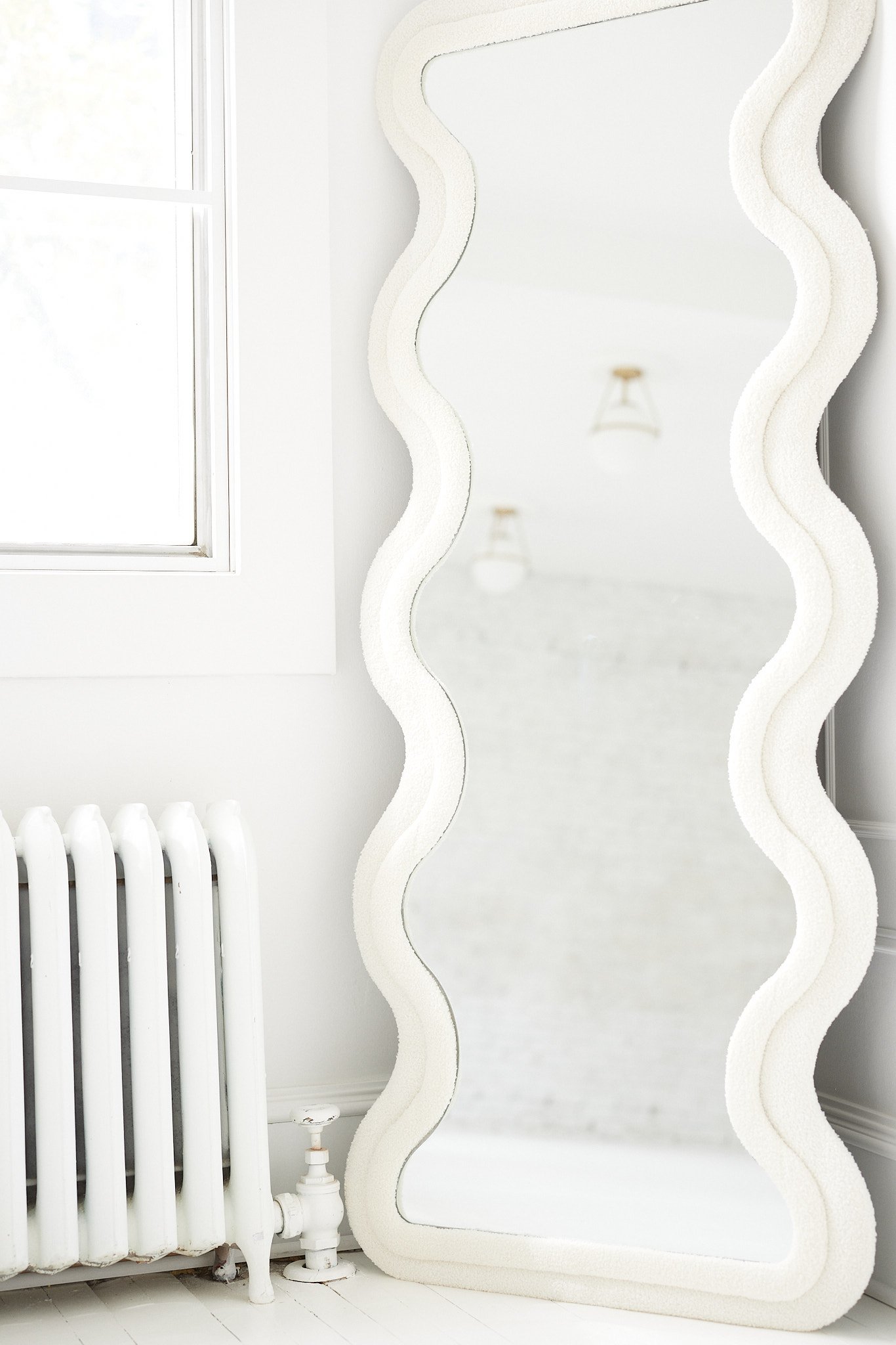 A white wavy-framed mirror leaning against a wall beside a white radiator, with a window and ceiling lights reflected in the mirror.